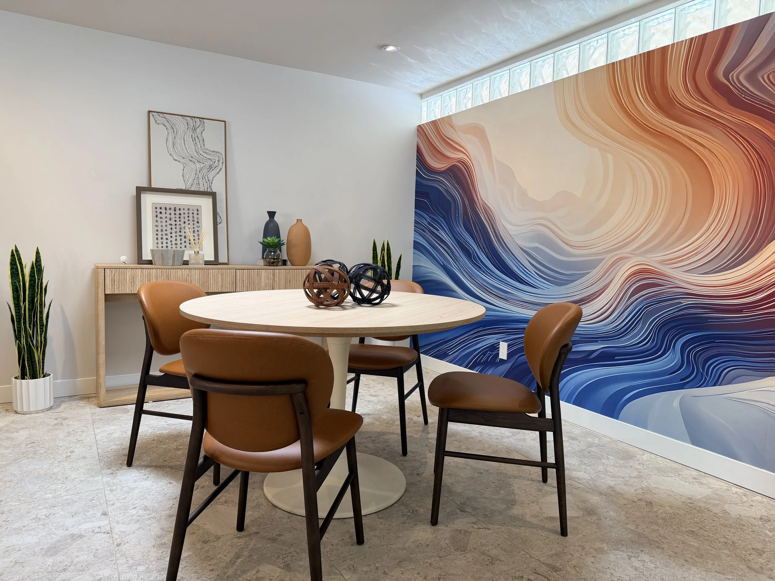 Interior of a dining room with a round white table, brown leather chairs, decorative black and brown geometric balls, a wooden sideboard with vases, artwork, and a potted plant. A large colorful wave mural decorates one wall, and a glass block window