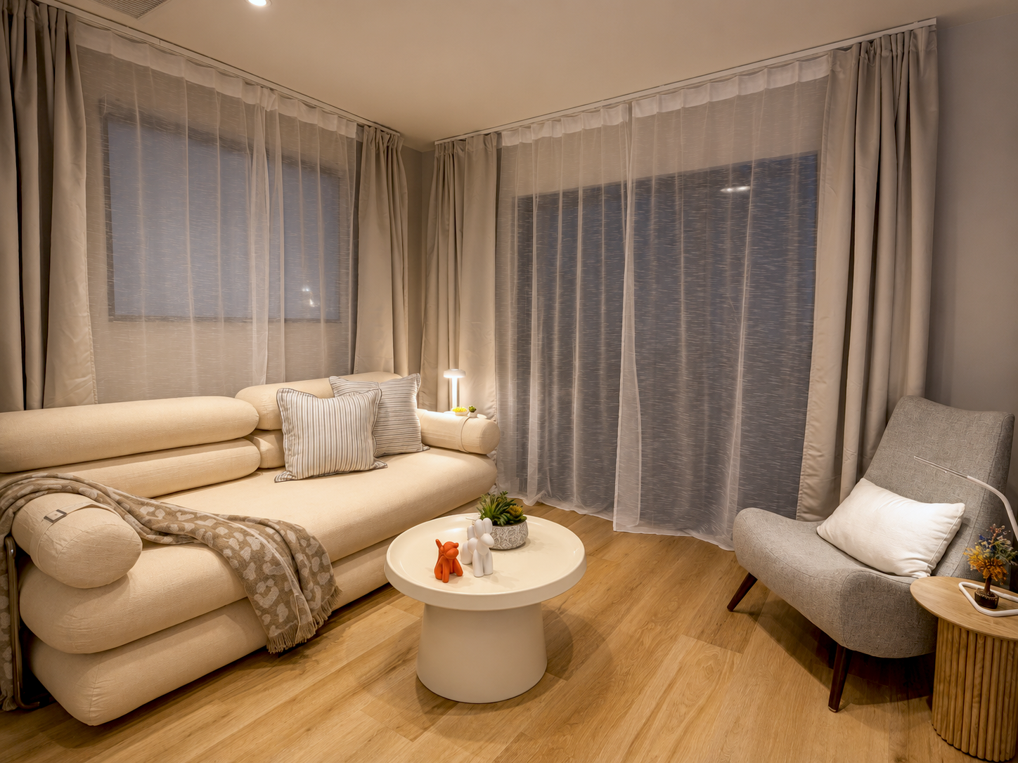 Living room with beige sofa, gray armchair, wooden floor, white round coffee table with decorative items, large windows with sheer curtains, side table with small pot of yellow flowers, small lamp, and pillows.