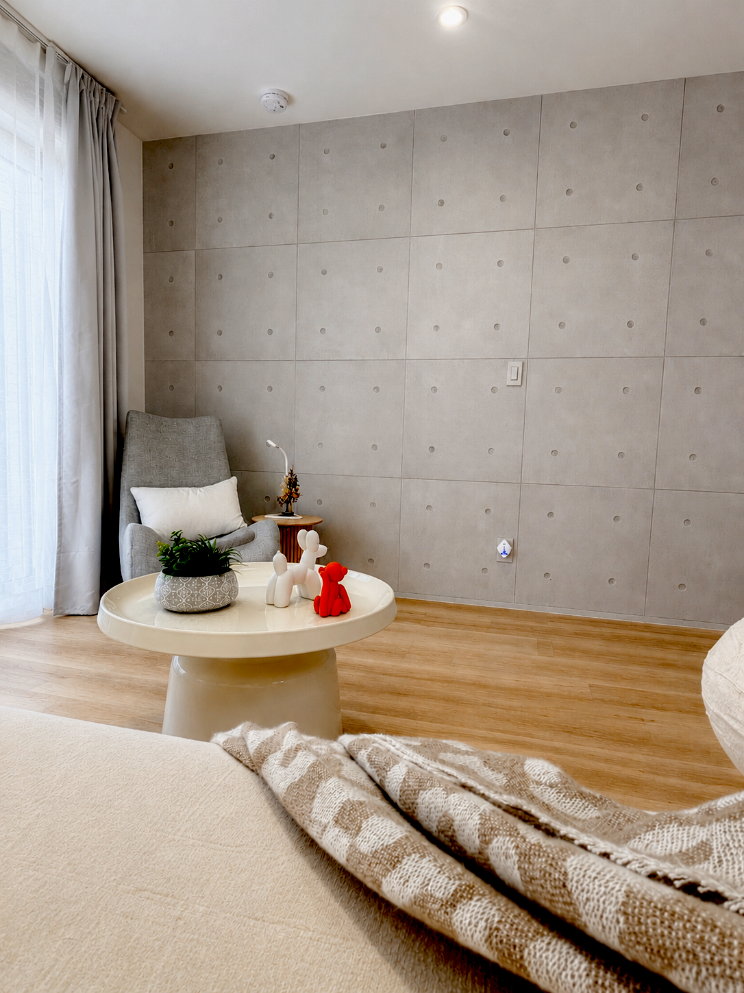 A cozy corner of a living room featuring a beige sofa with a patterned throw blanket. A white round coffee table displays small decorative toys, a potted plant, and miniature sculptures of dogs. A grey armchair with a white pillow and a small side ta