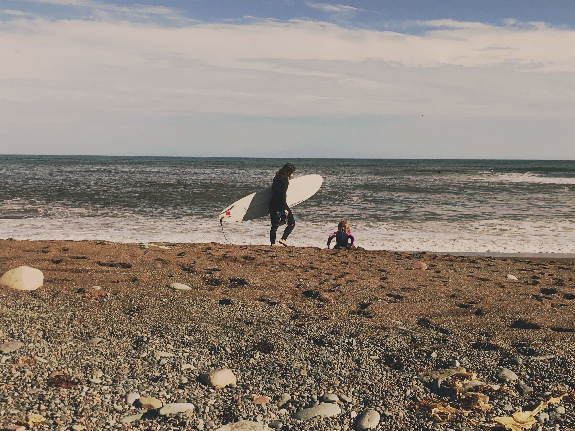 Woman holding a surfboard walking on a pebbled beach while a child sits on the shore, ocean waves in the background, cloudy sky overhead.