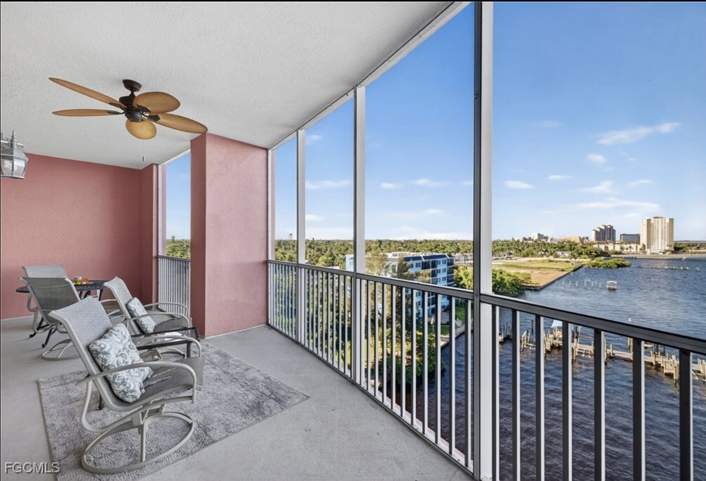 Balcony with beige chairs, a small table with drinks, pink wall, ceiling fan, and a view of a river, trees, and buildings under a blue sky.