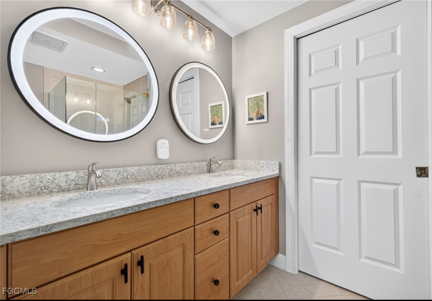 A bathroom vanity with a granite countertop, two undermount sinks, and wooden cabinets with black knobs. Two illuminated oval mirrors are mounted on the light gray wall above the vanity. To the right, there is a closed white panel door. A small framed picture hangs on the wall between the mirrors. The area reflected in the mirrors shows a shower with glass doors and a light fixture above.
