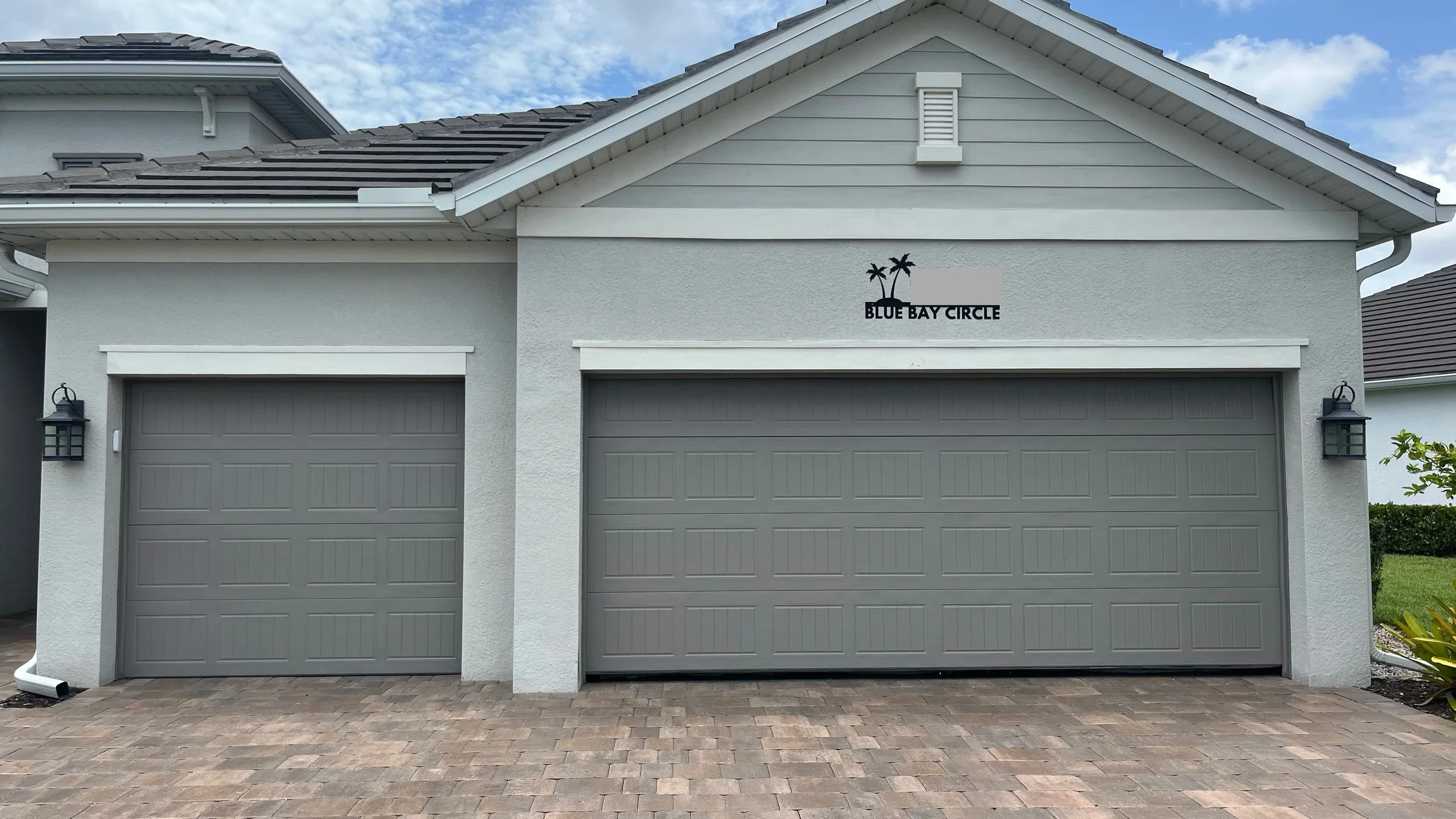 A modern house with two gray garage doors, exterior walls painted light gray, and a sign that reads 'Blue Bay Circle' with palm trees. Two black outdoor lanterns are mounted beside the garage doors, and the driveway is paved with brick.
