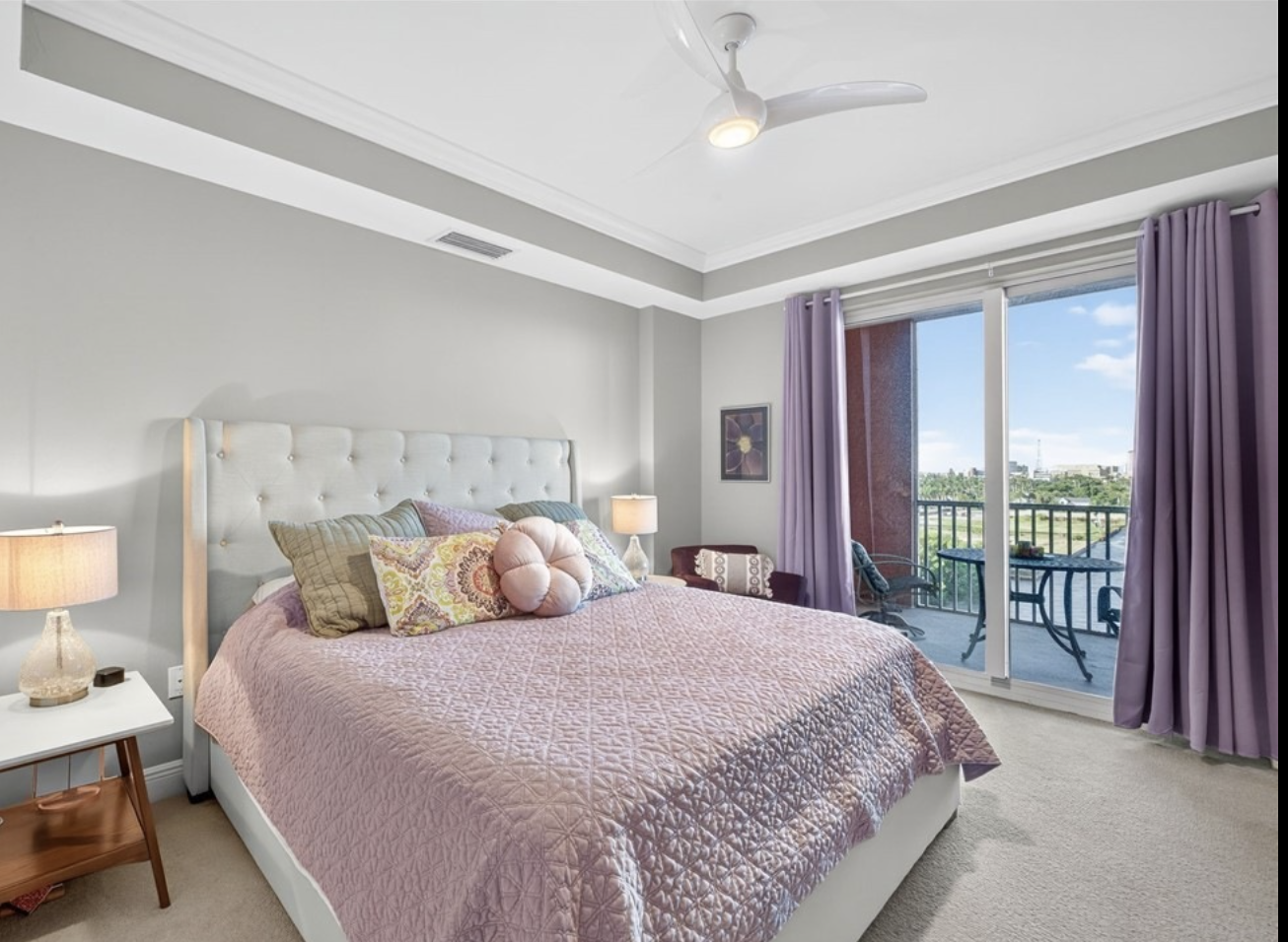 Bedroom with a large bed with a pink quilt and multiple pillows, nightstands with lamps, a balcony with outdoor furniture, and purple curtains.