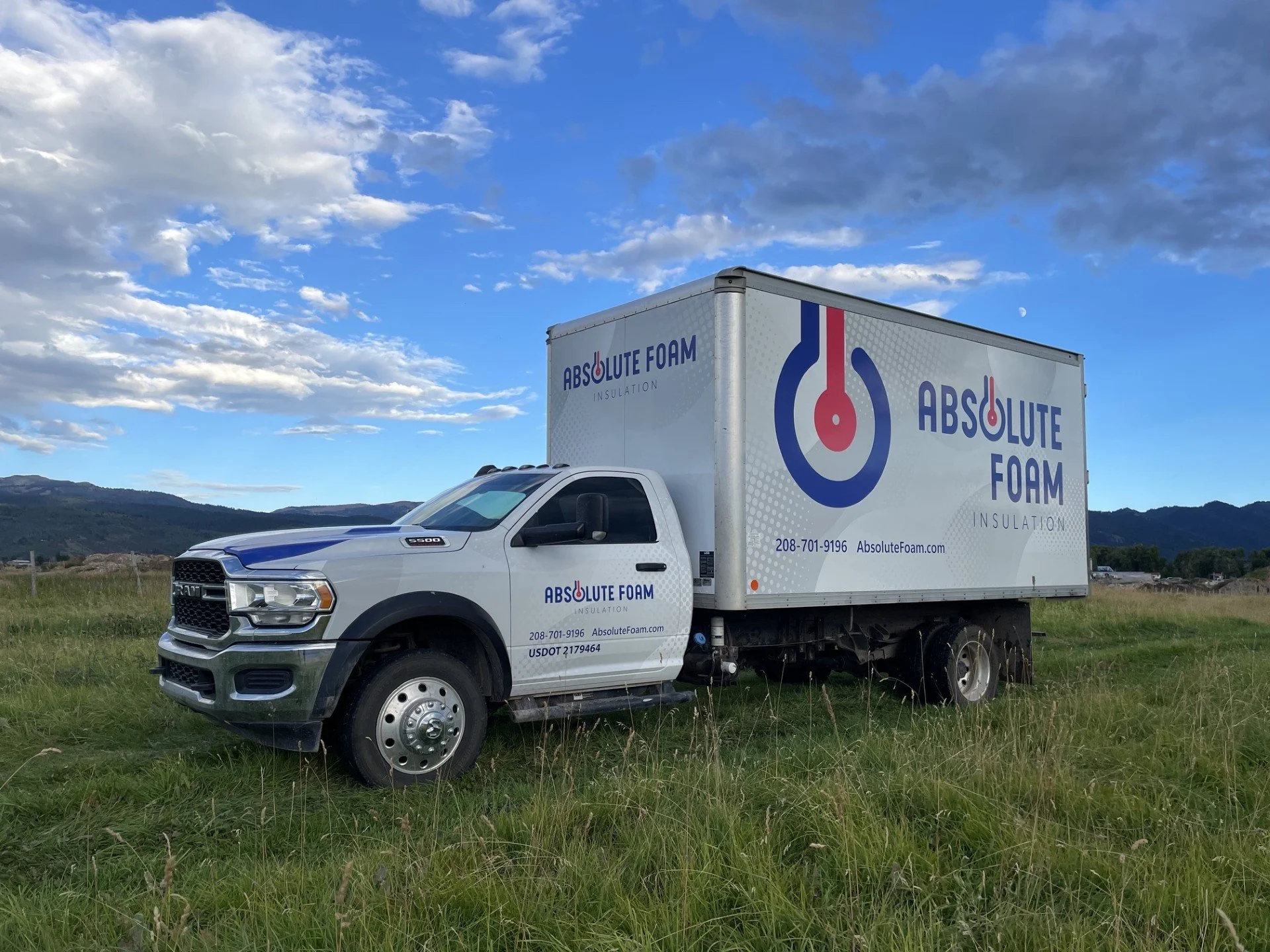 A white truck with a large cargo area labeled 'Absolute Foam Insulation' parked on a grassy field with mountains in the background. The sky is partly cloudy with the moon visible.