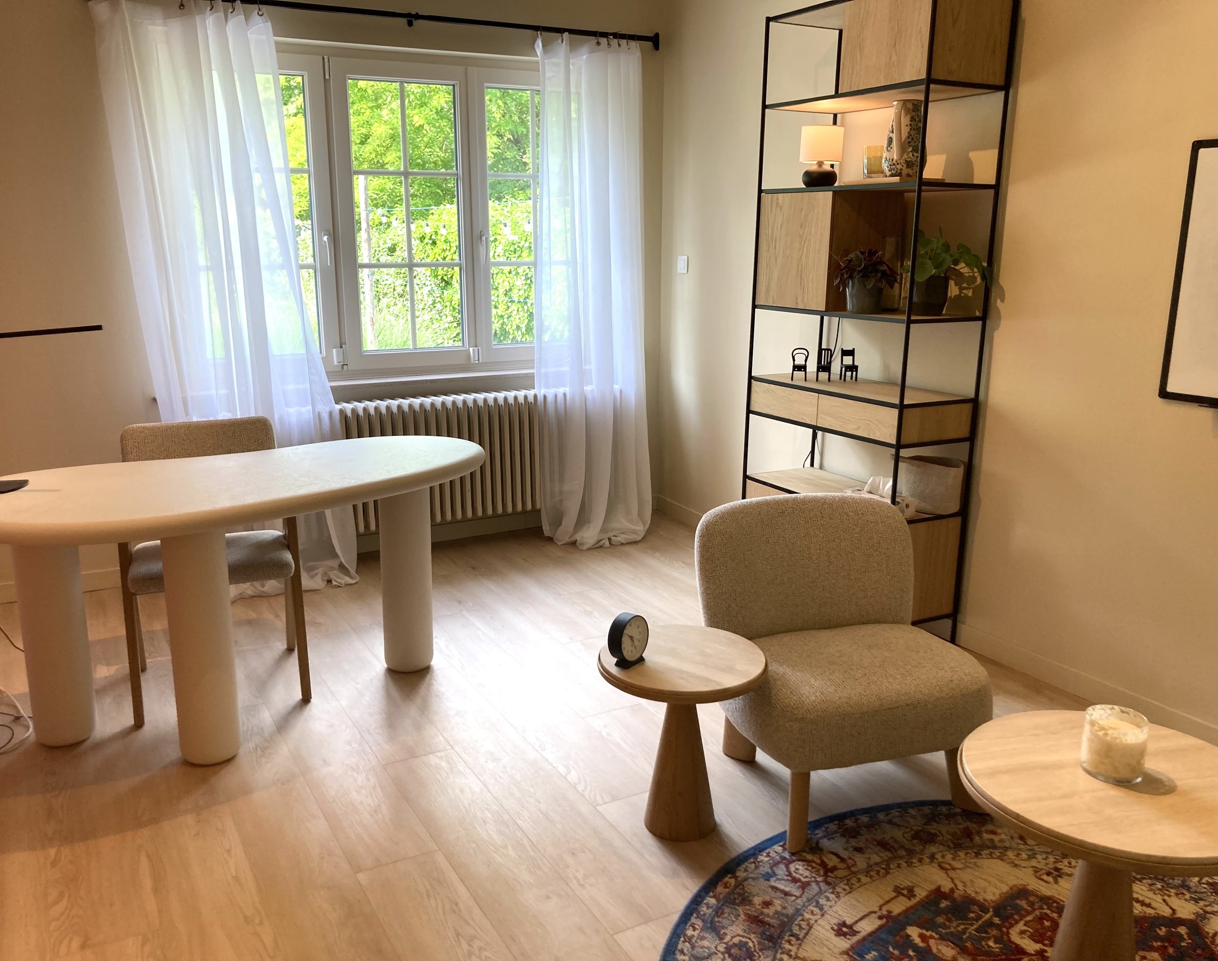 Een woonkamer met grote ramen met witte gordijnen, houten vloer, en een wandrek met decoratieve items zoals lampen en planten.