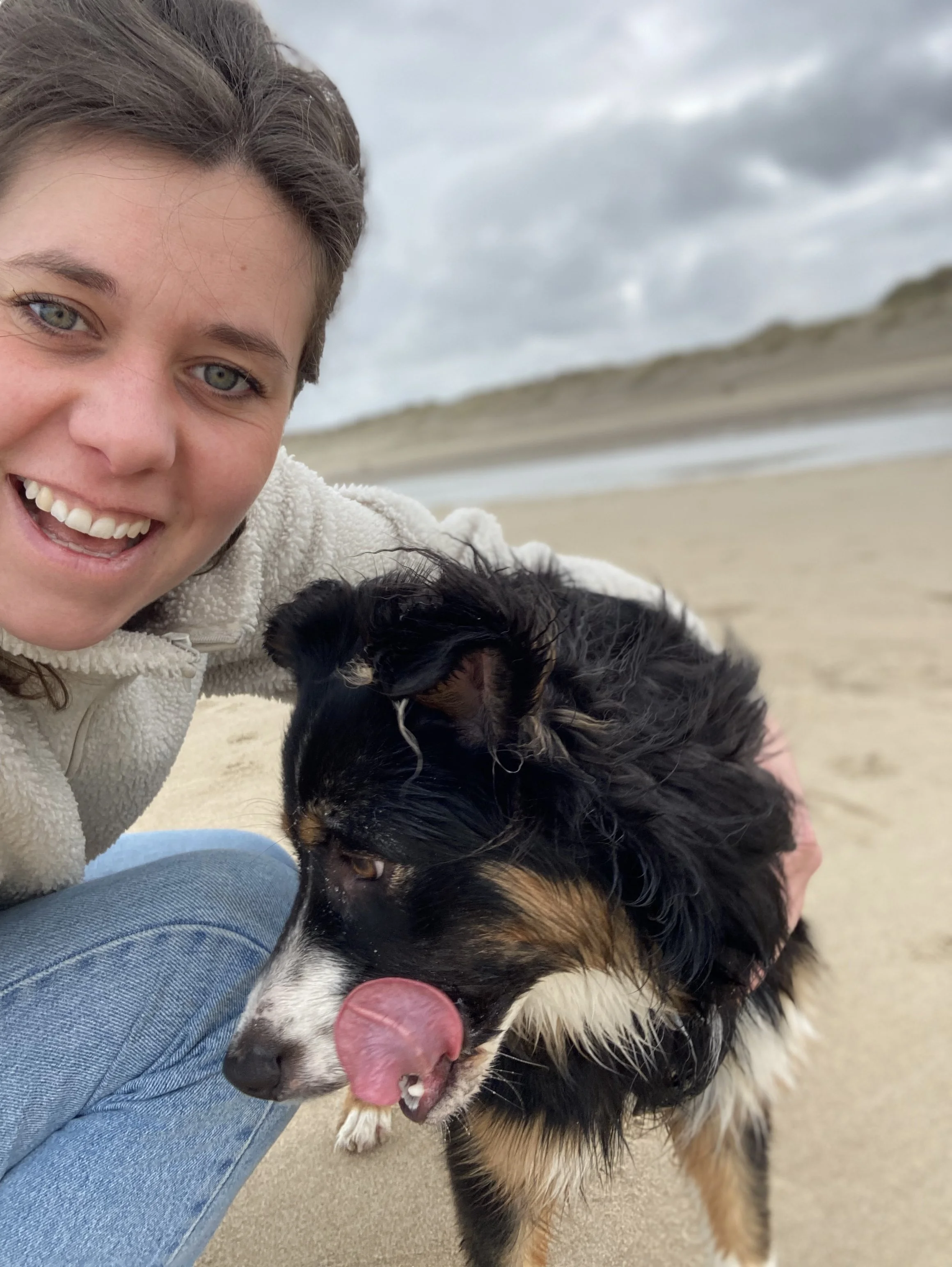Een glimlachende vrouw op het strand met haar hond. De achtergrond toont een bewolkte hemel en duinen.