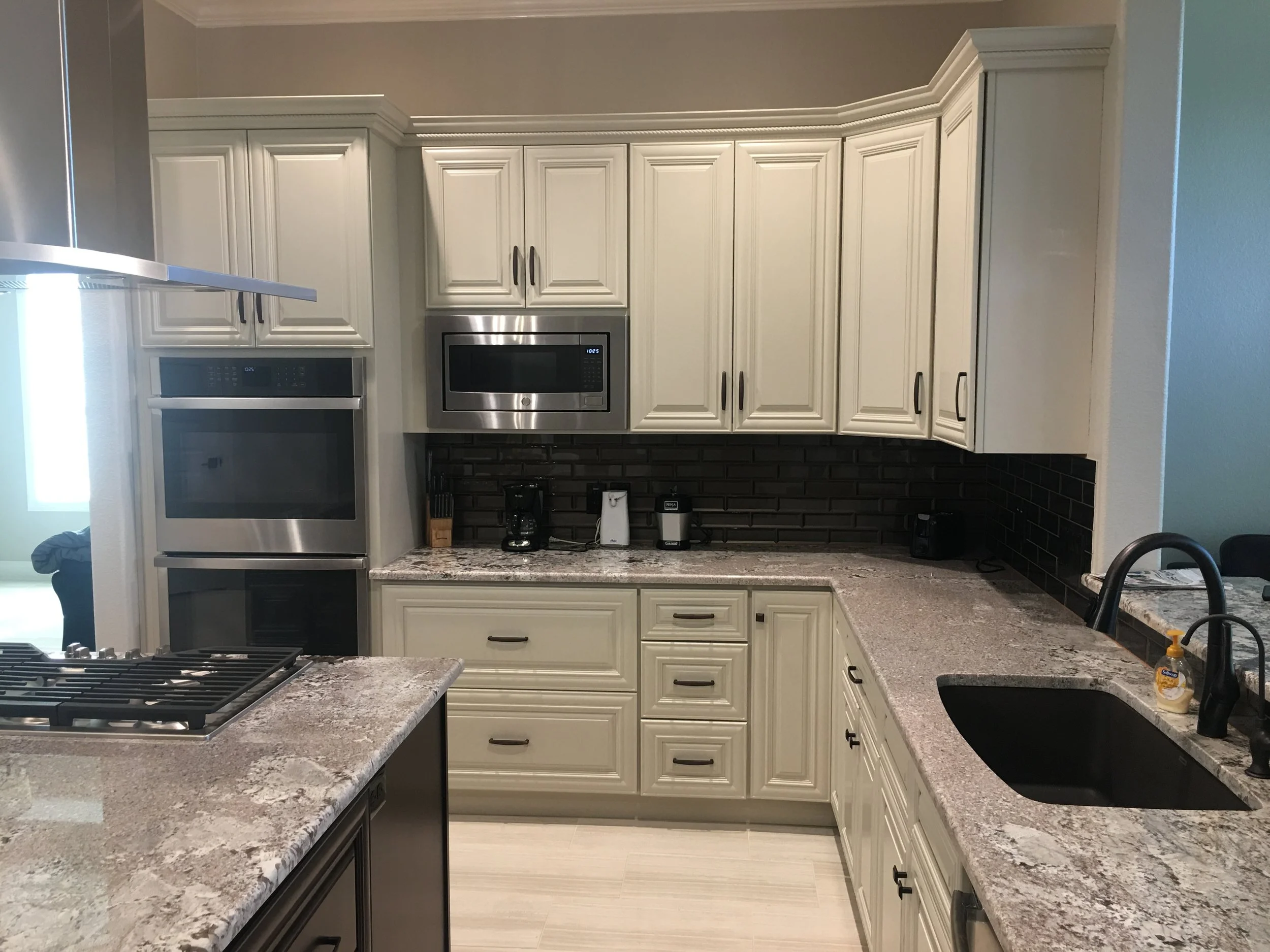 A modern kitchen featuring white cabinetry with dark handles, granite countertops, and black subway tile backsplash. Stainless steel appliances include a microwave and oven, with a gas stove in the foreground. There are items such as a coffee maker a