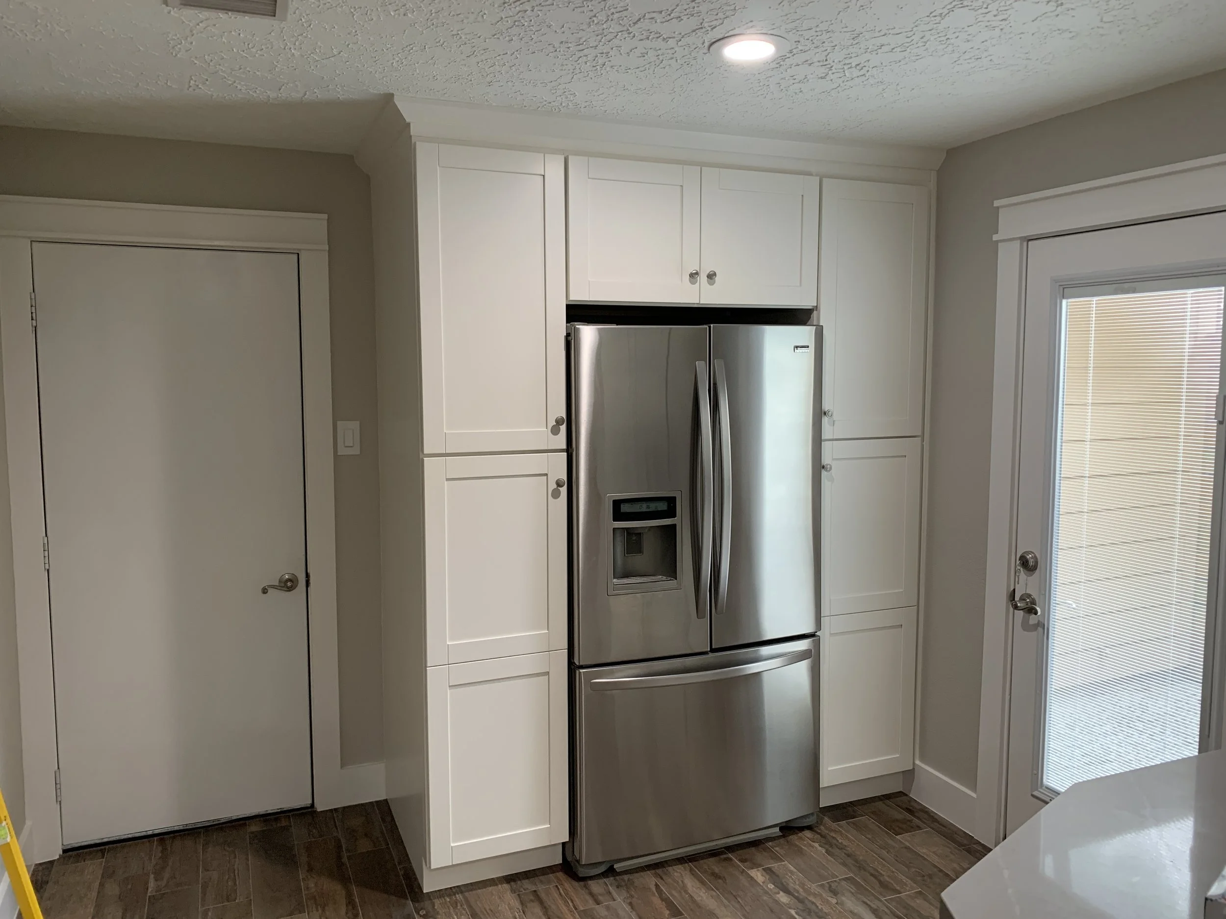 A kitchen with white cabinets, a stainless steel refrigerator, a window with blinds, and a textured ceiling.