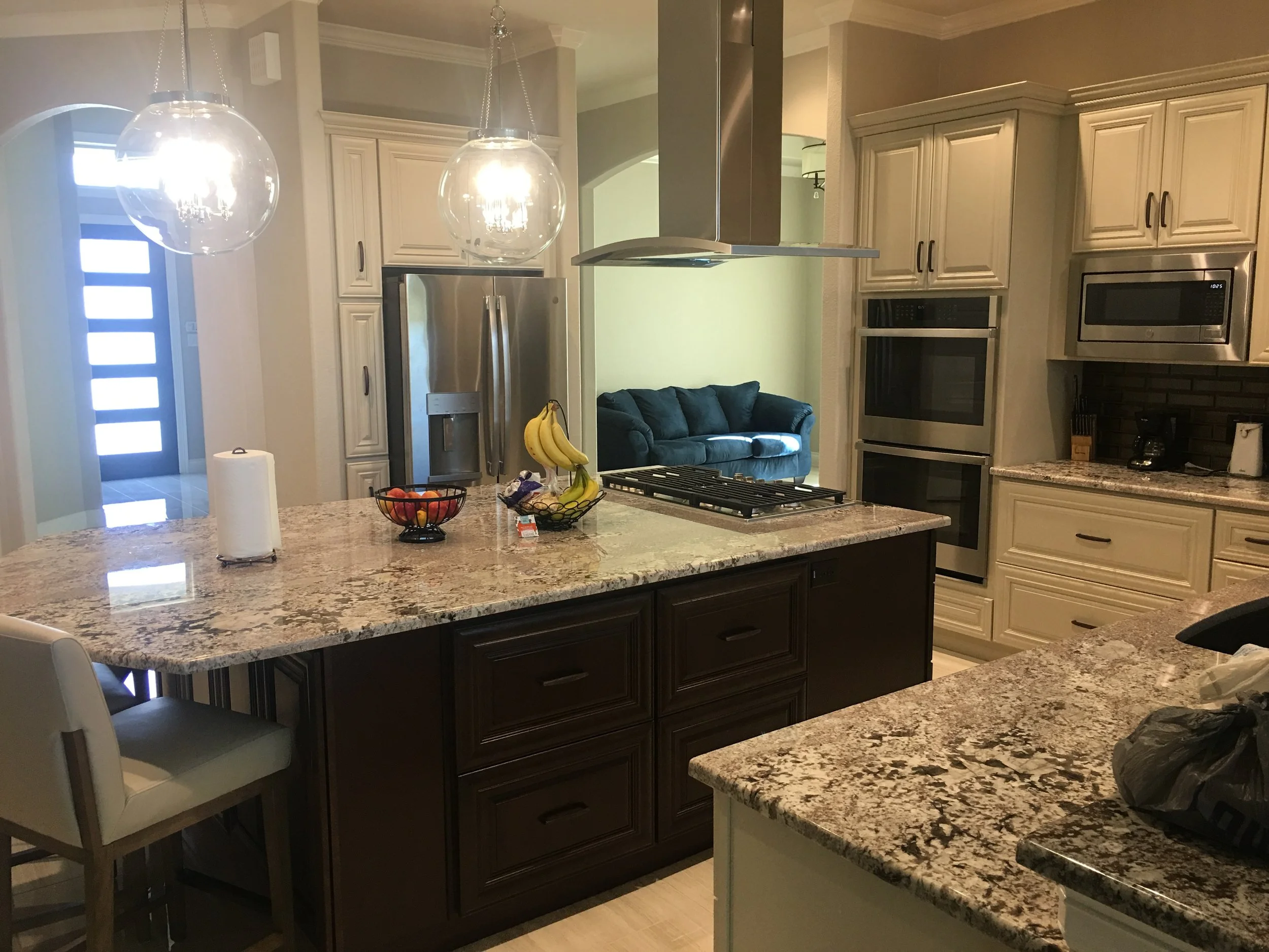 Kitchen with granite countertops, light-colored cabinetry, stainless steel appliances including a microwave and double oven. Two pendant lights hang above the island, which has a fruit basket and paper towel roll on it. In the background, a living ro