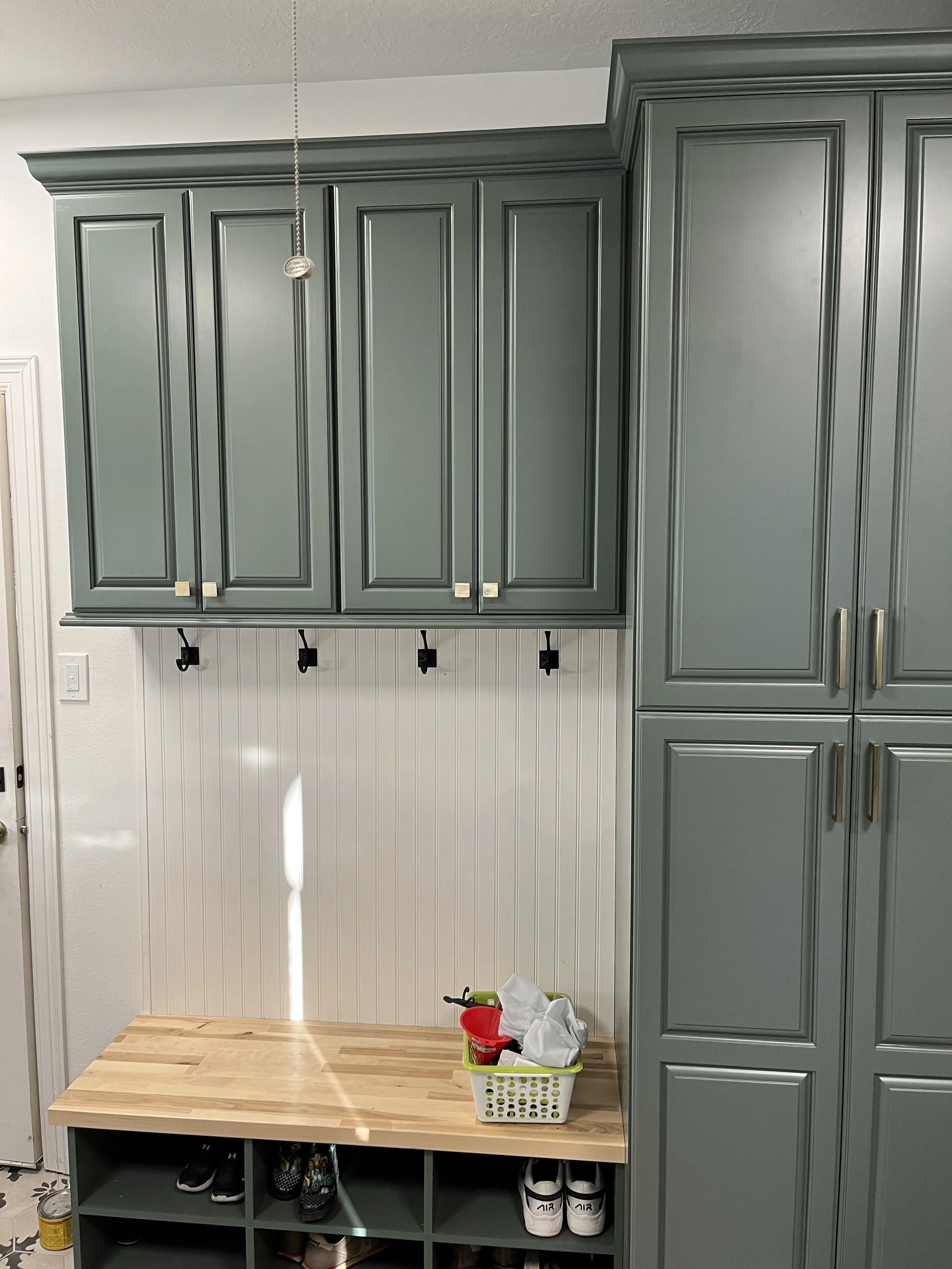 Living room area with gray built-in cabinets, a small wooden bench, and a shoe storage cubby underneath. There is a basket with some items on top of the bench, and hooks on the wall below the cabinets. Part of a white wall with beadboard paneling can