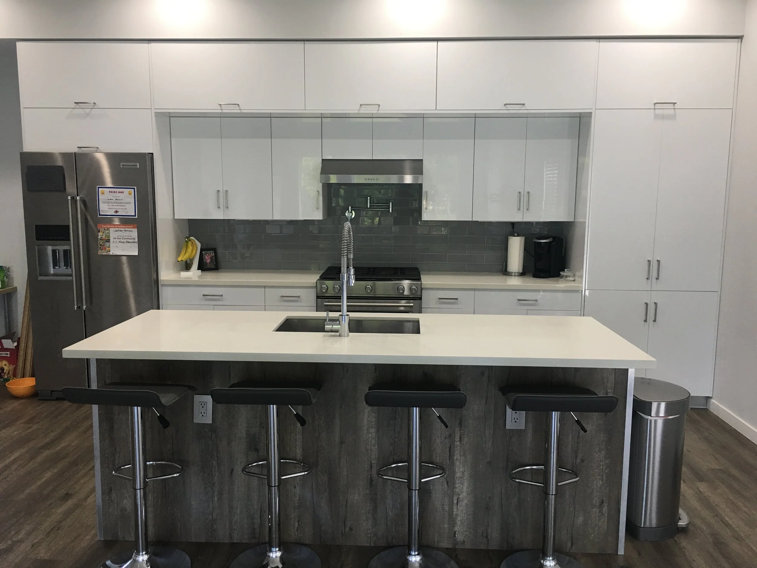 A modern kitchen with white cabinets, a stainless steel refrigerator, a gray tiled backsplash, and a kitchen island with a sink and three black bar stools.