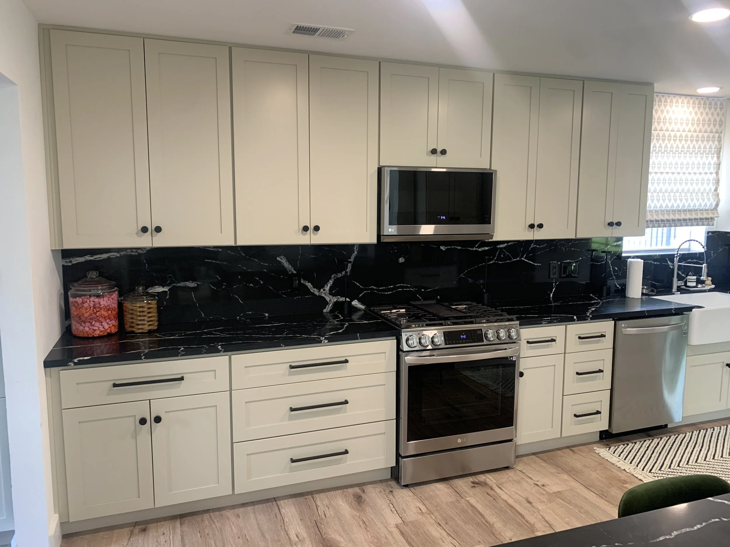 Kitchen with cream-colored cabinets, black handles, black marble countertop, stainless steel LG microwave, stove, and oven, near a window with patterned blinds, and a striped rug on wooden floor.