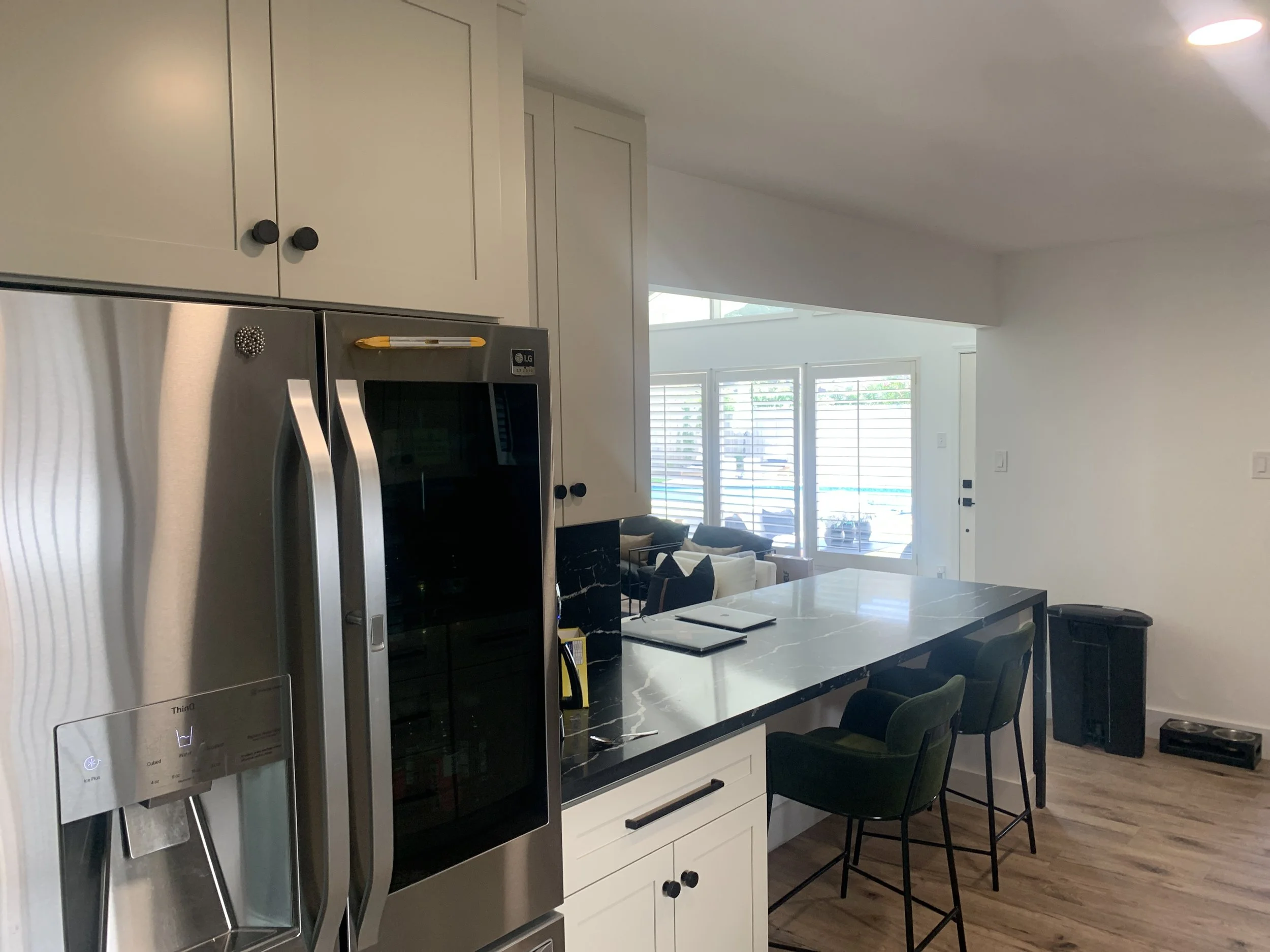 Modern kitchen and dining area with white cabinets, stainless steel refrigerator, marble countertop island, and black chairs. Large windows with blinds allow natural light to fill the space.