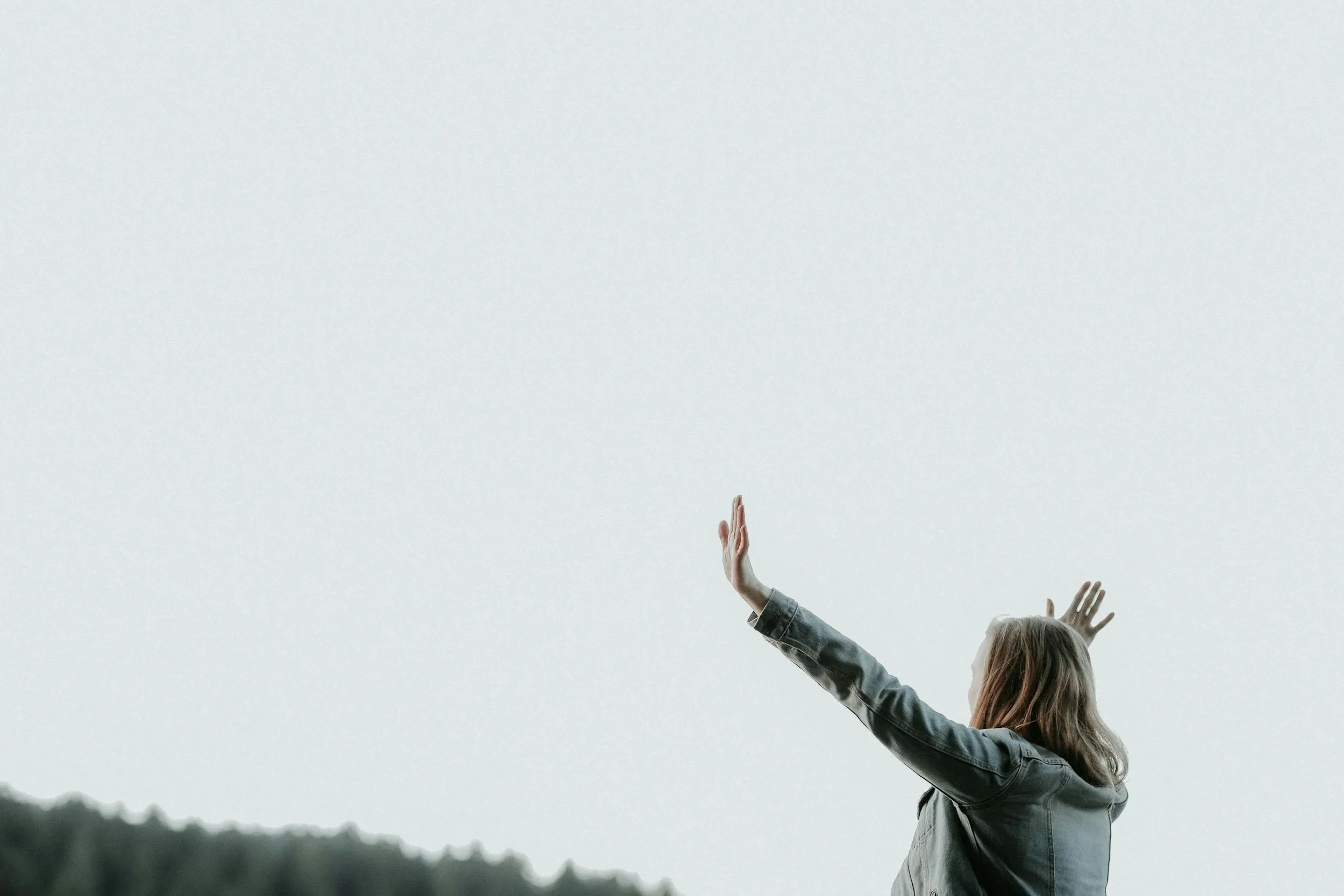 A woman with long hair wearing a denim jacket raising her arms in front of an overcast sky, with mountains and trees in the background.