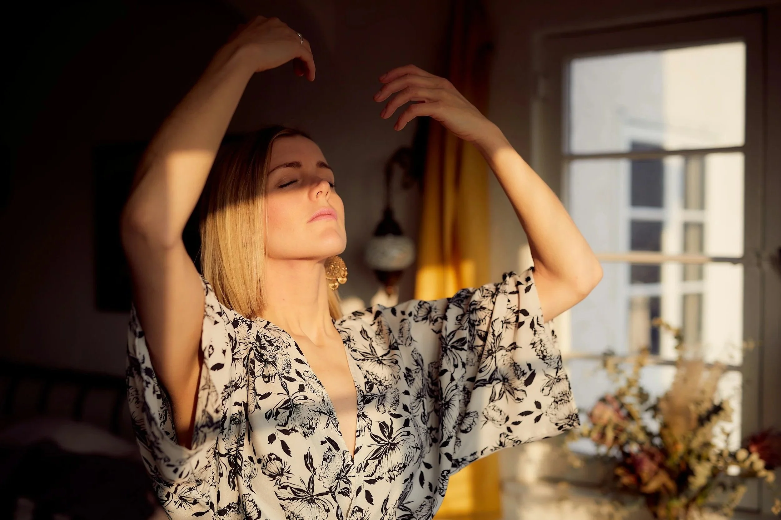 A woman with blonde hair and closed eyes, wearing a black and white floral dress, is standing with her arms raised and hands curved above her head in a peaceful pose, near a window with sunlight streaming in.
