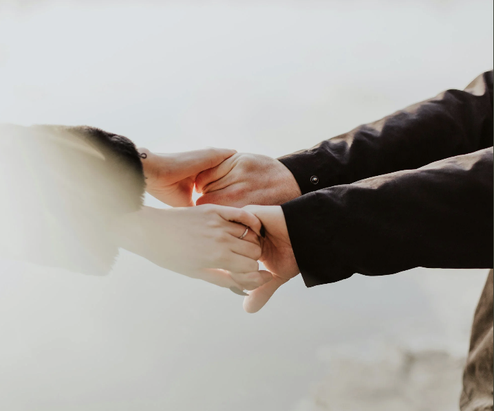 Two people holding hands, one with a ring, against a cloudy sky background