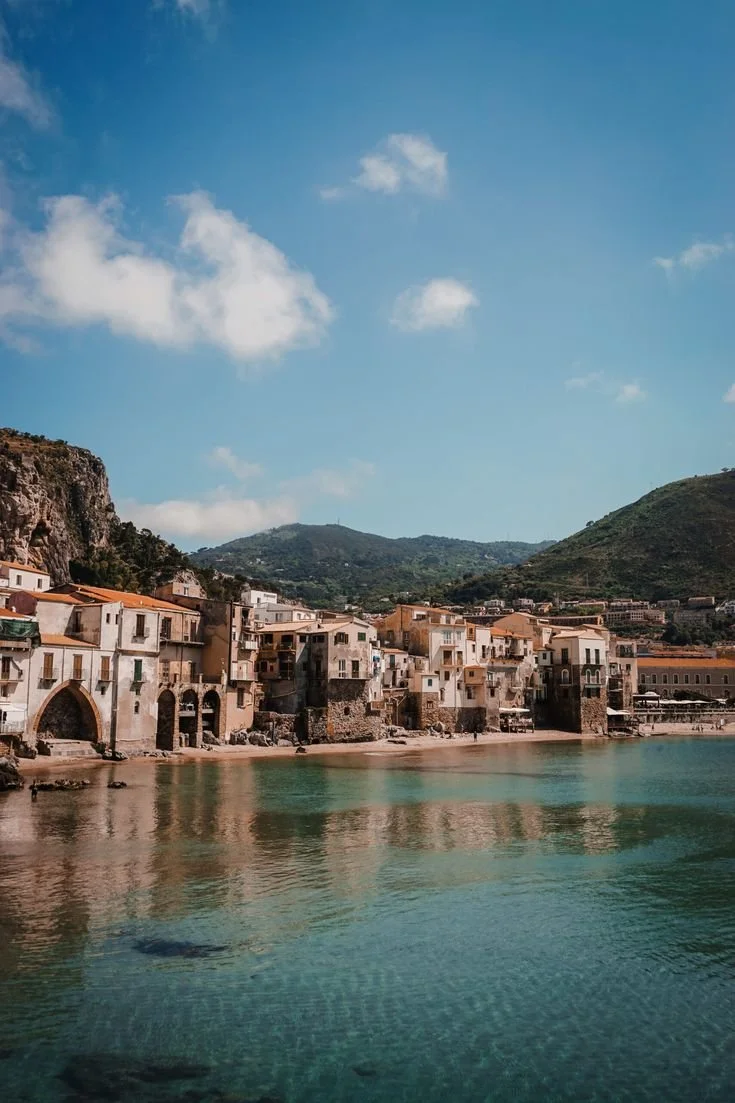 Coastal town with colorful buildings next to turquoise water and green hills nearby under a blue sky with a few white clouds.