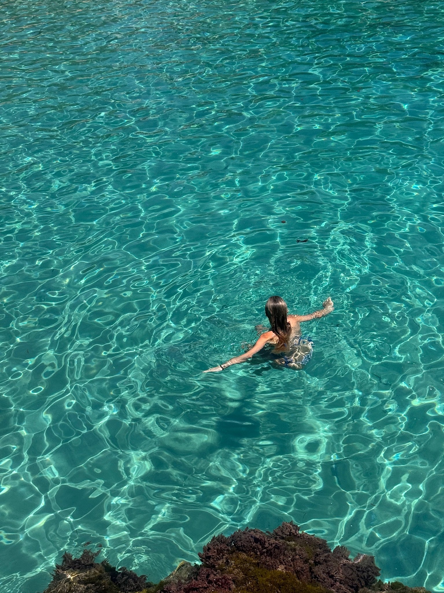 A person with long hair swimming in clear, turquoise water near rocky shoreline.