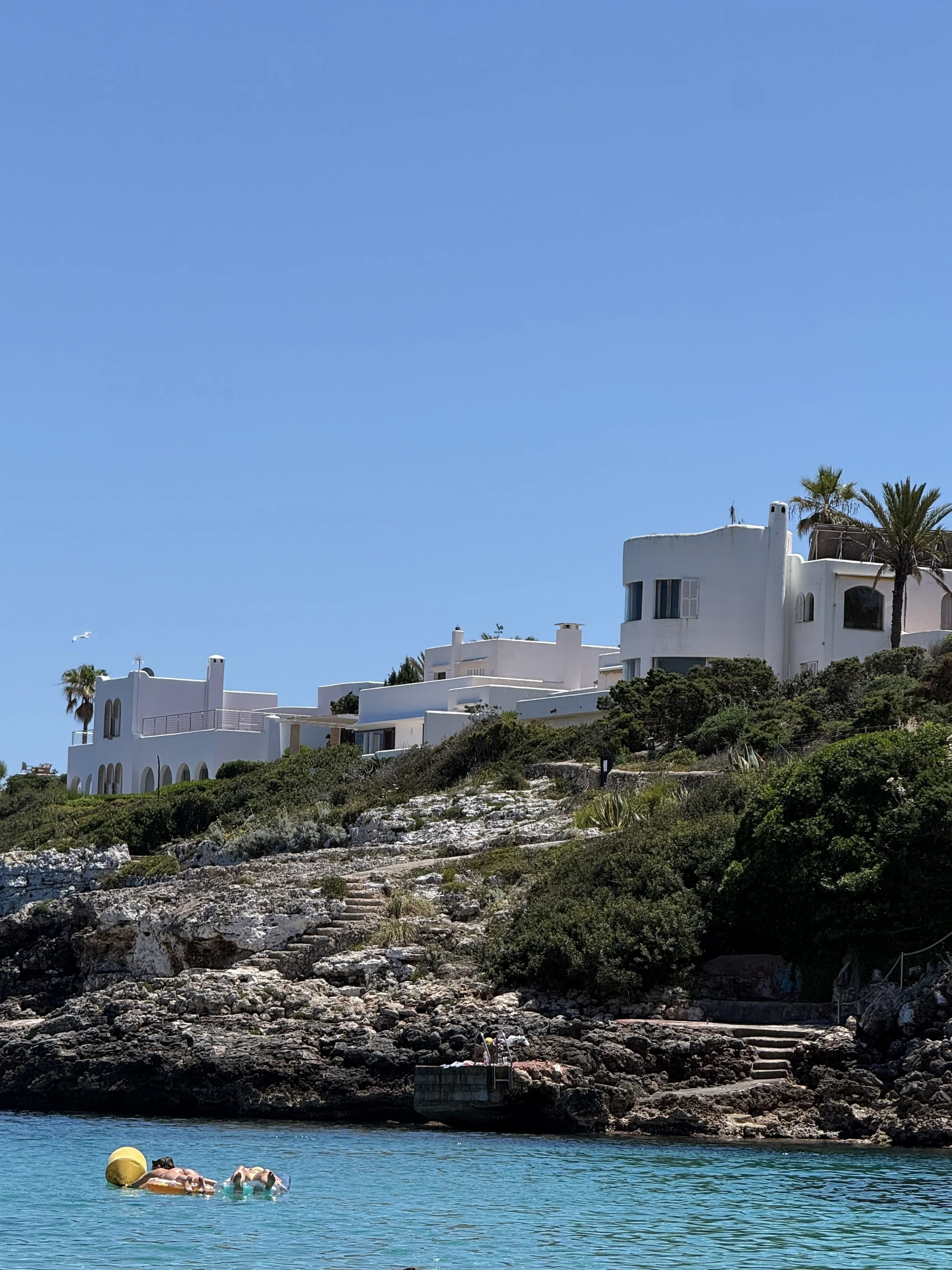 People relaxing on a float in clear blue water near rocky shoreline with white buildings and palm trees on hill in background under a bright blue sky