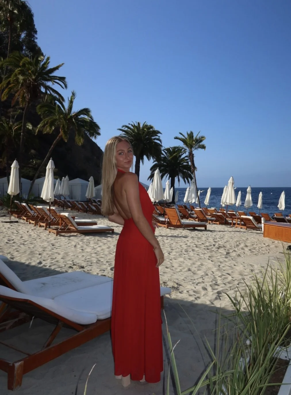 A woman in a red dress standing on a sandy beach with palm trees, lounge chairs, and white umbrellas, overlooking the ocean under a clear blue sky.