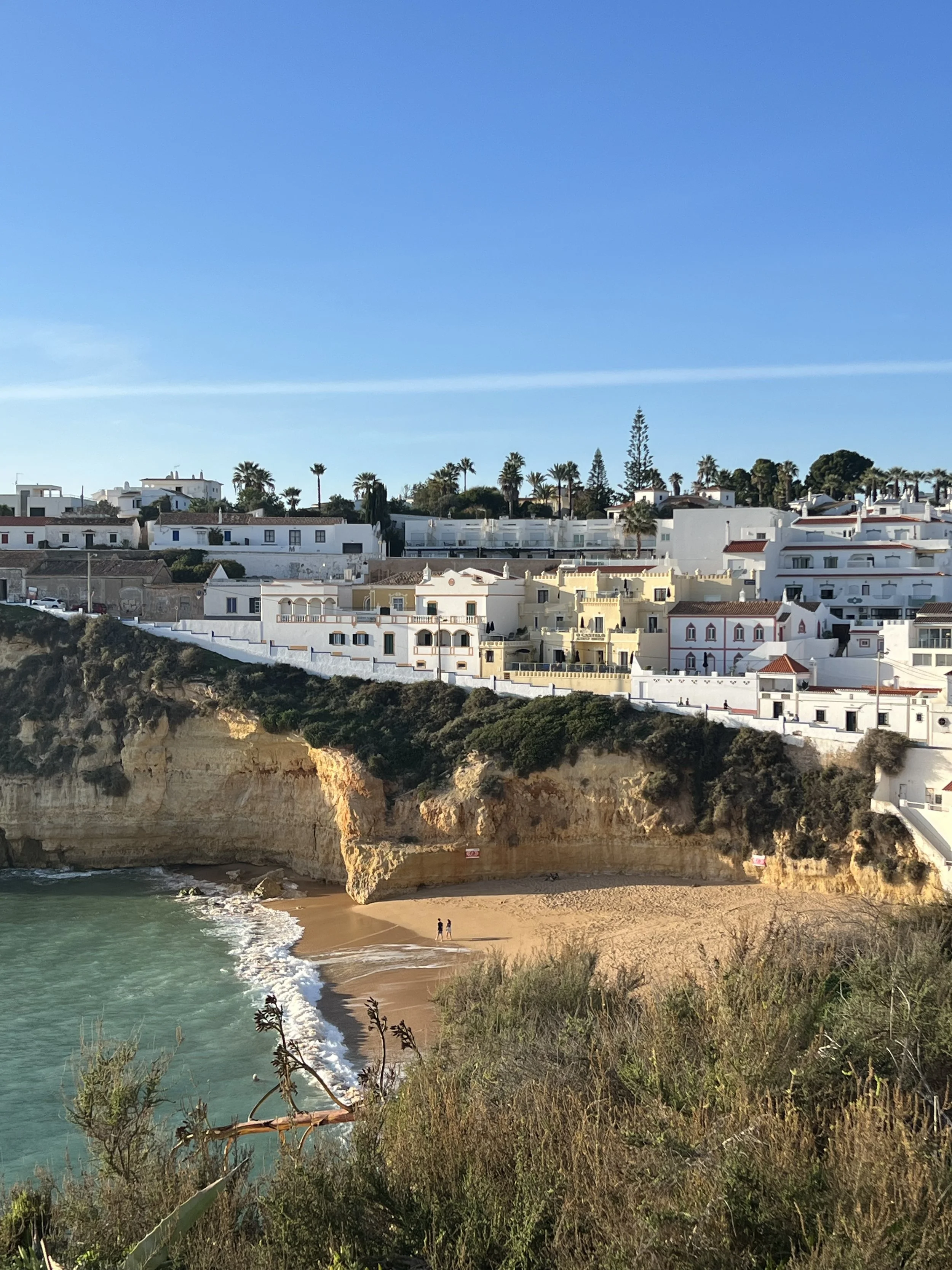View of a beach with golden sand, turquoise water, cliffs, and white buildings with red roofs and palm trees on a hillside.