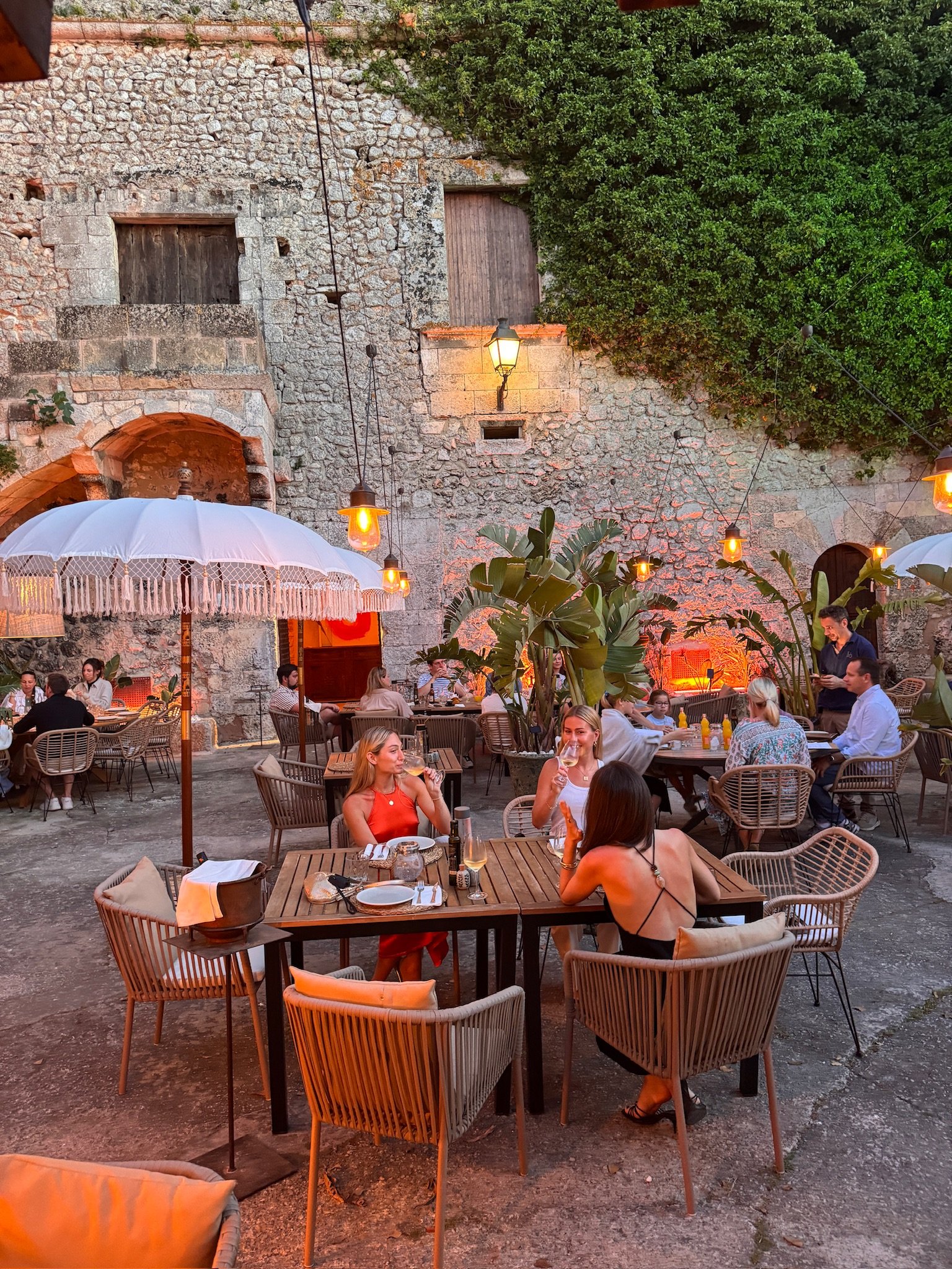 People dining outdoors at a restaurant with rustic stone walls, greenery, and decorative string lights and lamps hanging from the ceiling in the evening.