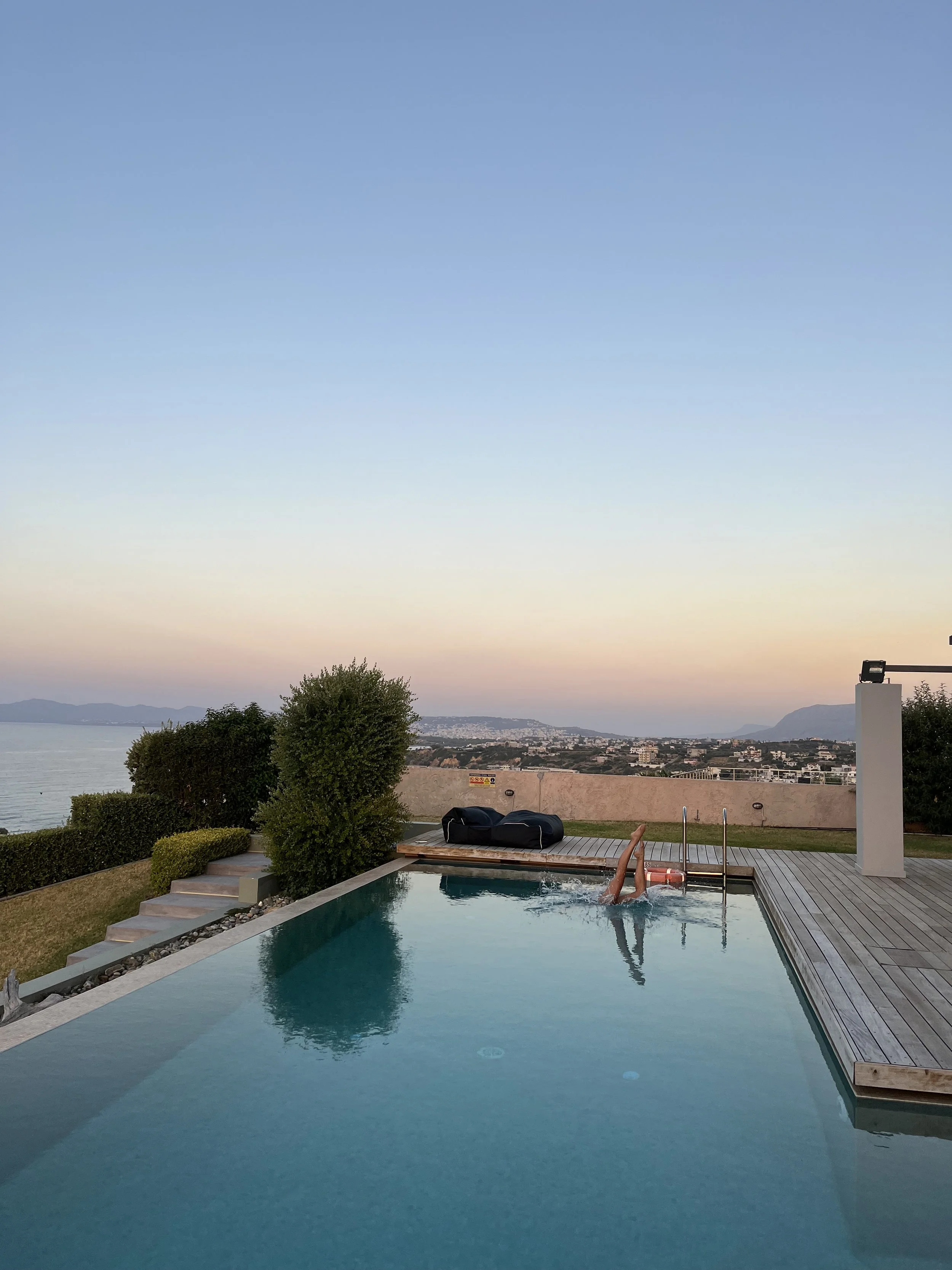 Person swimming in a backyard pool with a city and mountains view at sunset.