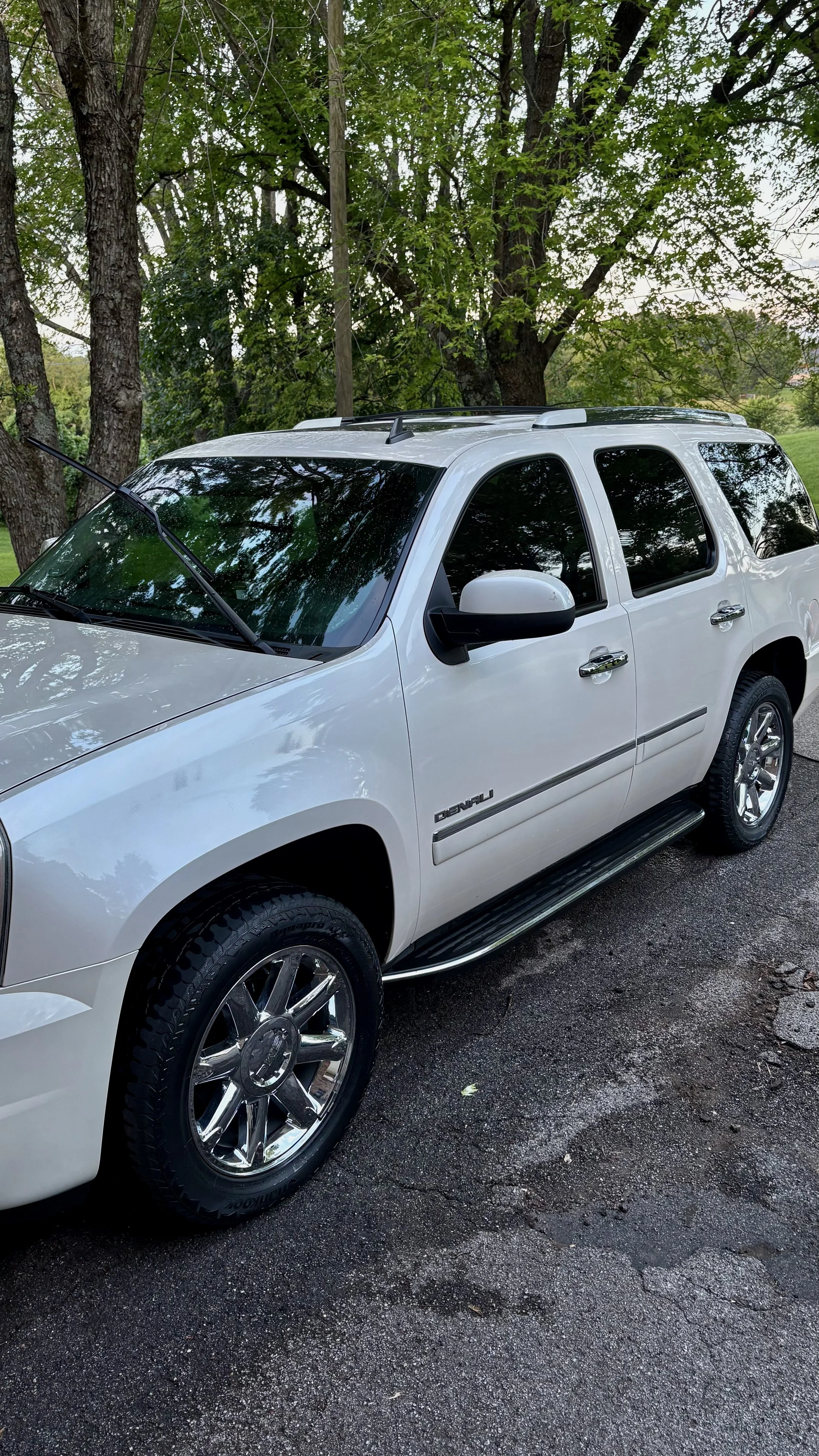 A white GMC Denali truck parked on a paved surface with trees and greenery in the background.