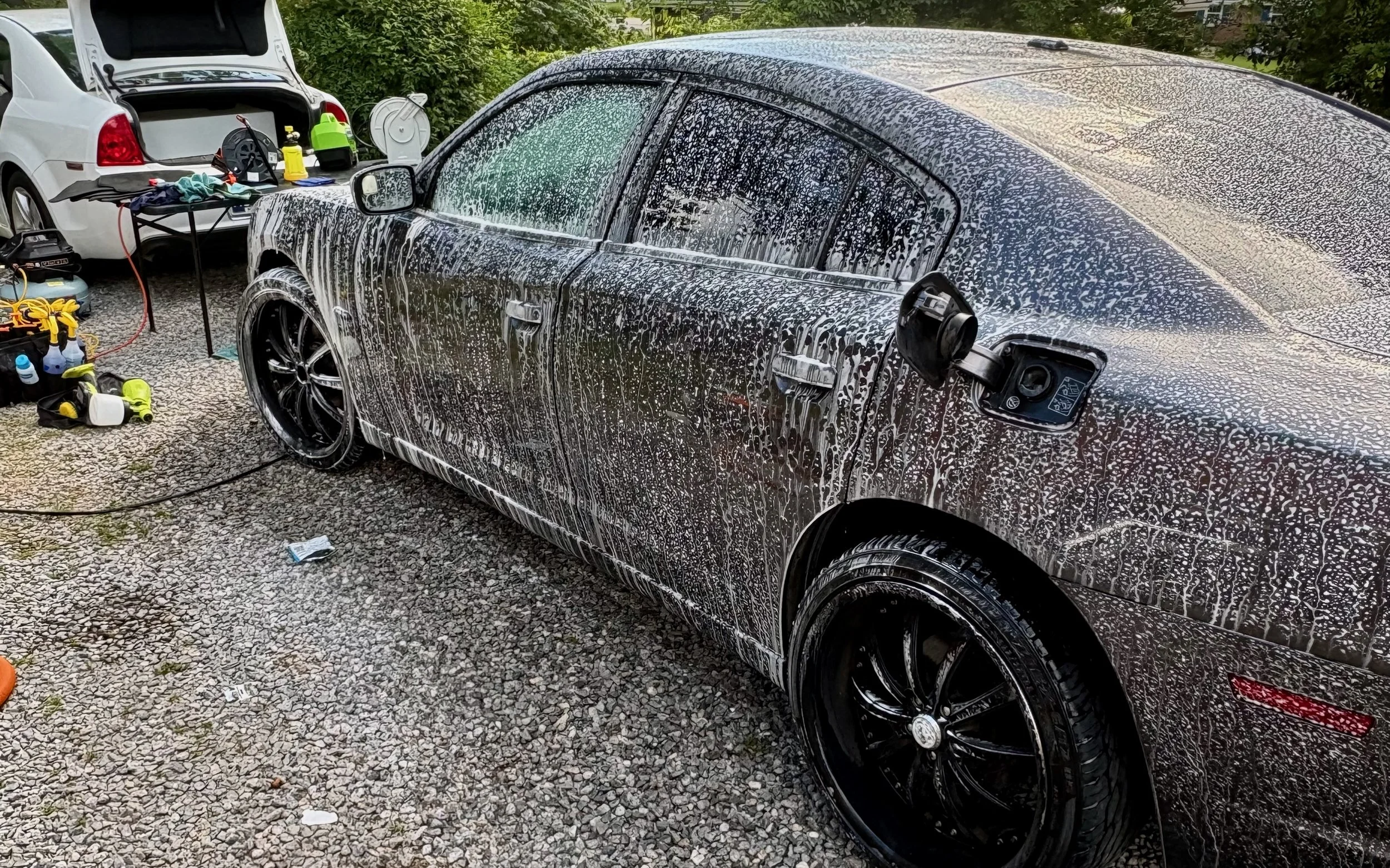 A black car being washed with soap and water, with foam covering the surface, parked on a gravel driveway. Nearby, there is a white car with its trunk open, and various car cleaning tools and supplies are visible.
