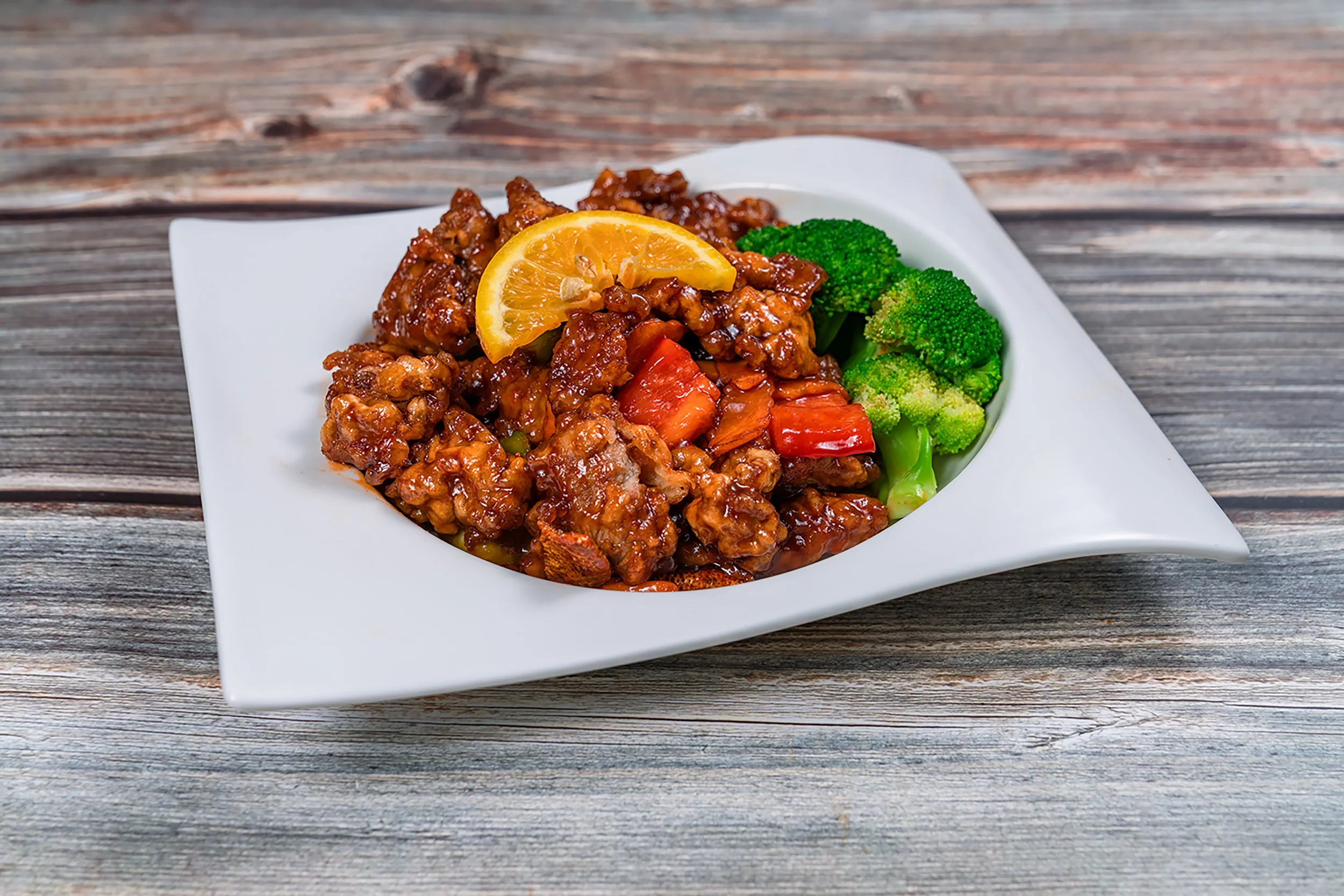 Close-up of a white square bowl containing sweet and sour chicken with a lemon slice on top, served with broccoli and red peppers, on a wooden table.
