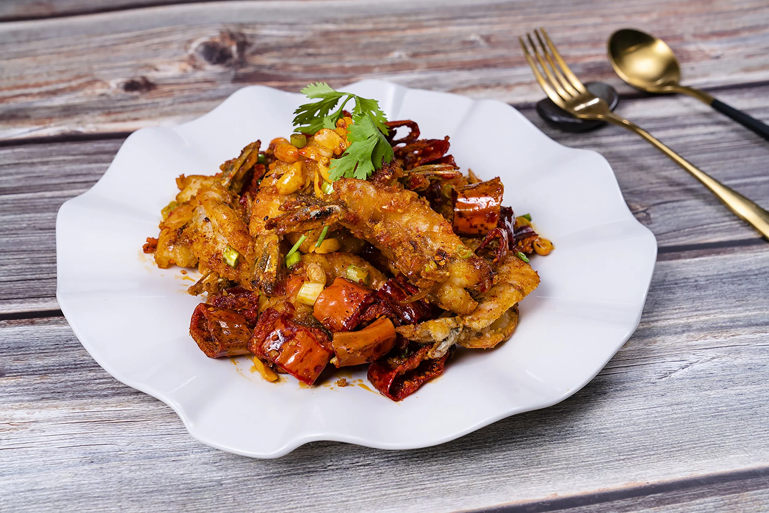 A white plate with a spicy seafood dish garnished with cilantro on a rustic wooden table, with a gold fork and spoon nearby.