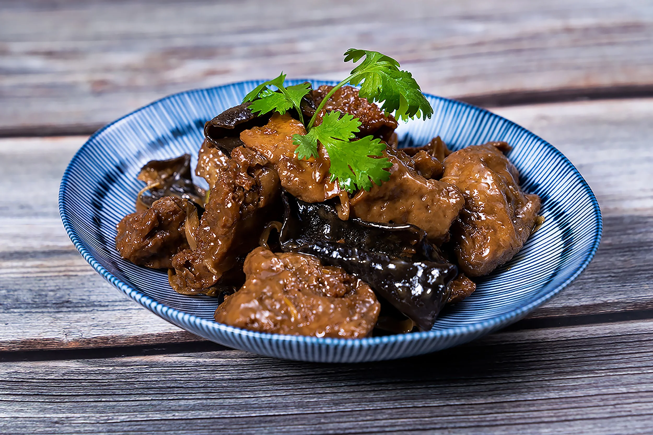 A blue-striped bowl containing braised meat and eggplant garnished with cilantro, placed on a weathered wooden surface.