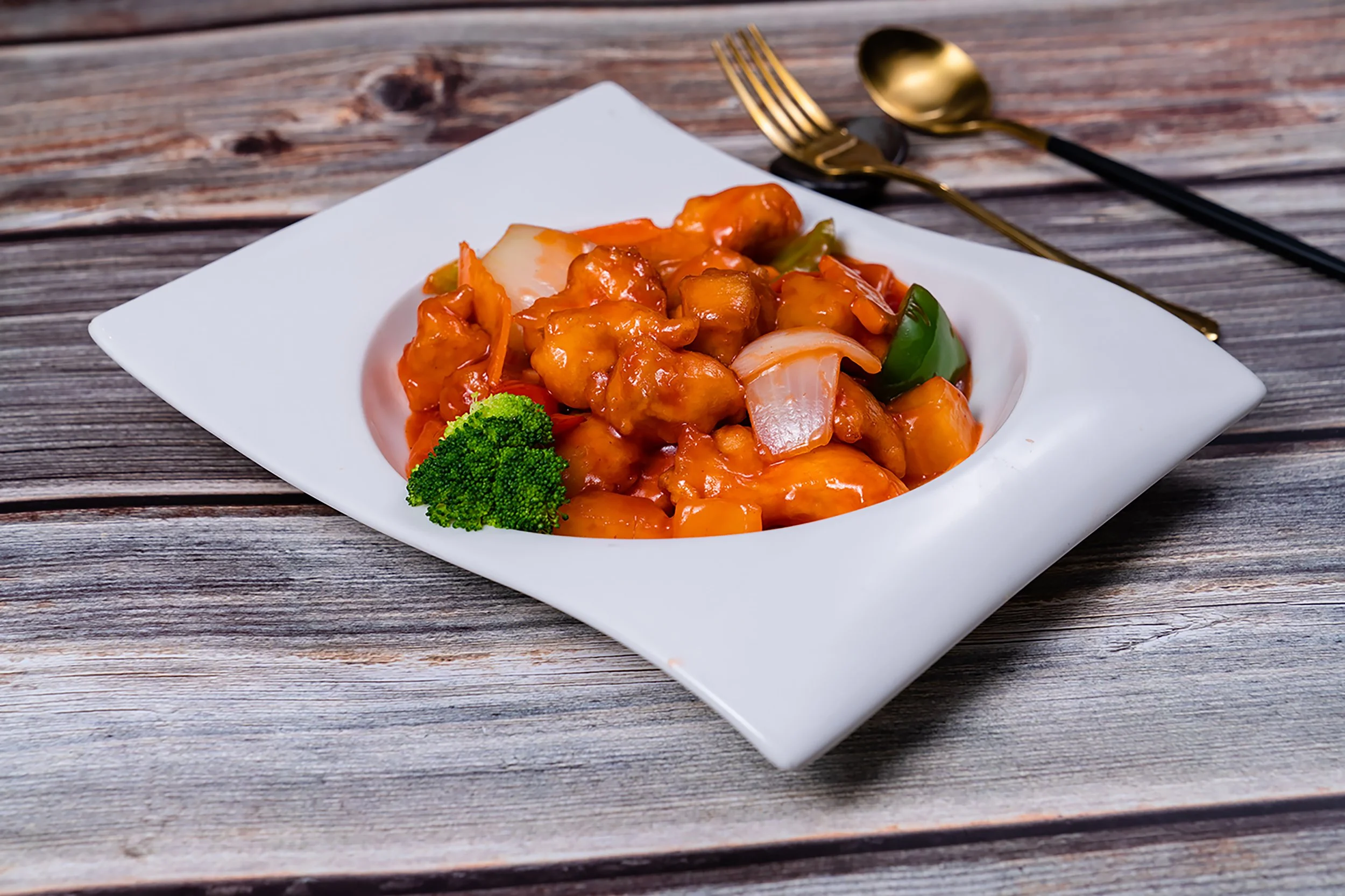 A white rectangular plate with sweet and sour chicken topped with orange sauce, accompanied by broccoli and green bell peppers, placed on a rustic wooden table with gold-colored fork and spoon in the background.