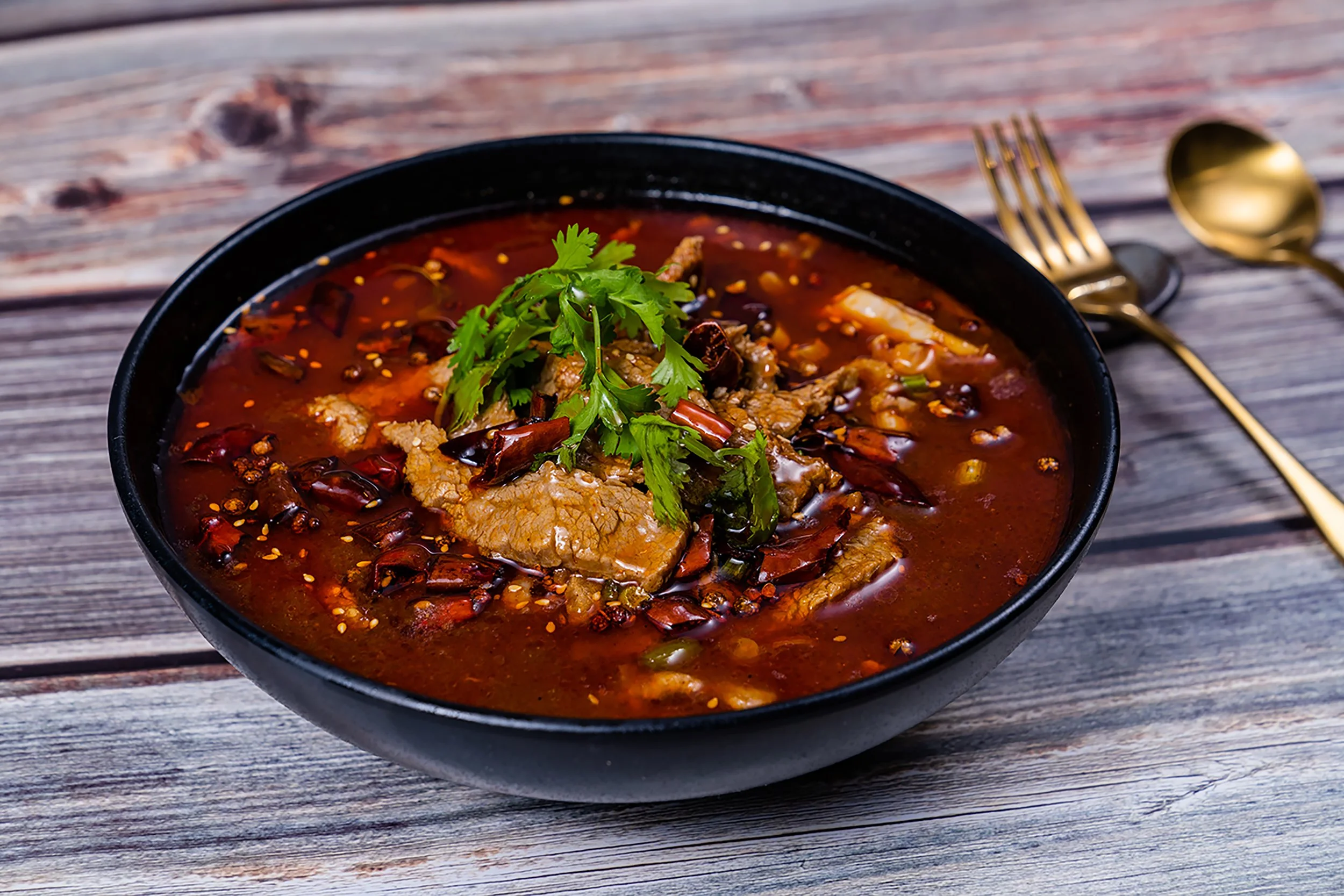 A black bowl of spicy Chinese hot pot soup garnished with fresh cilantro, sitting on a rustic wooden table with gold-colored spoon and fork.