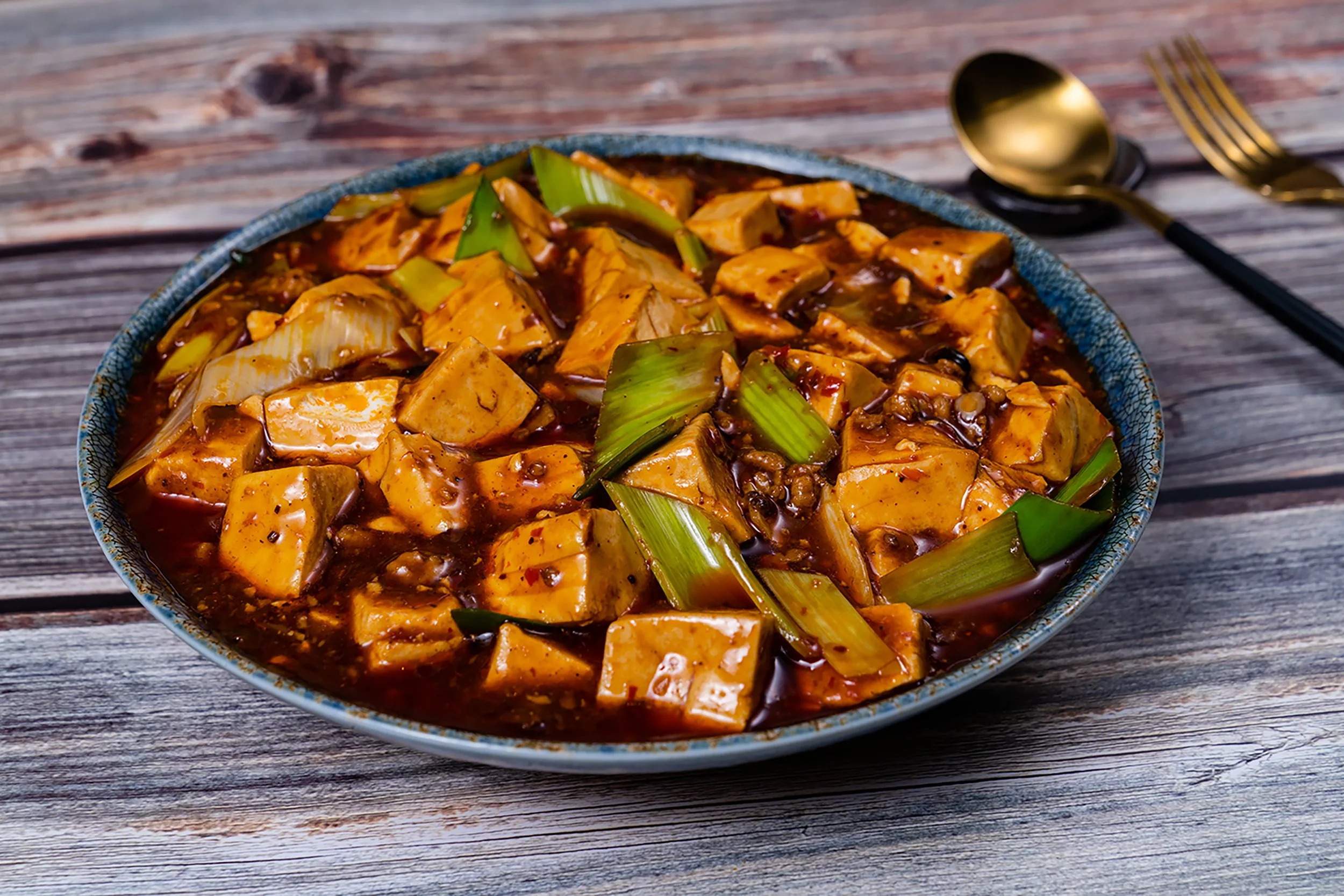 A bowl of spicy tofu and vegetable stir-fry in a dark sauce, garnished with green onions, on a wooden table.