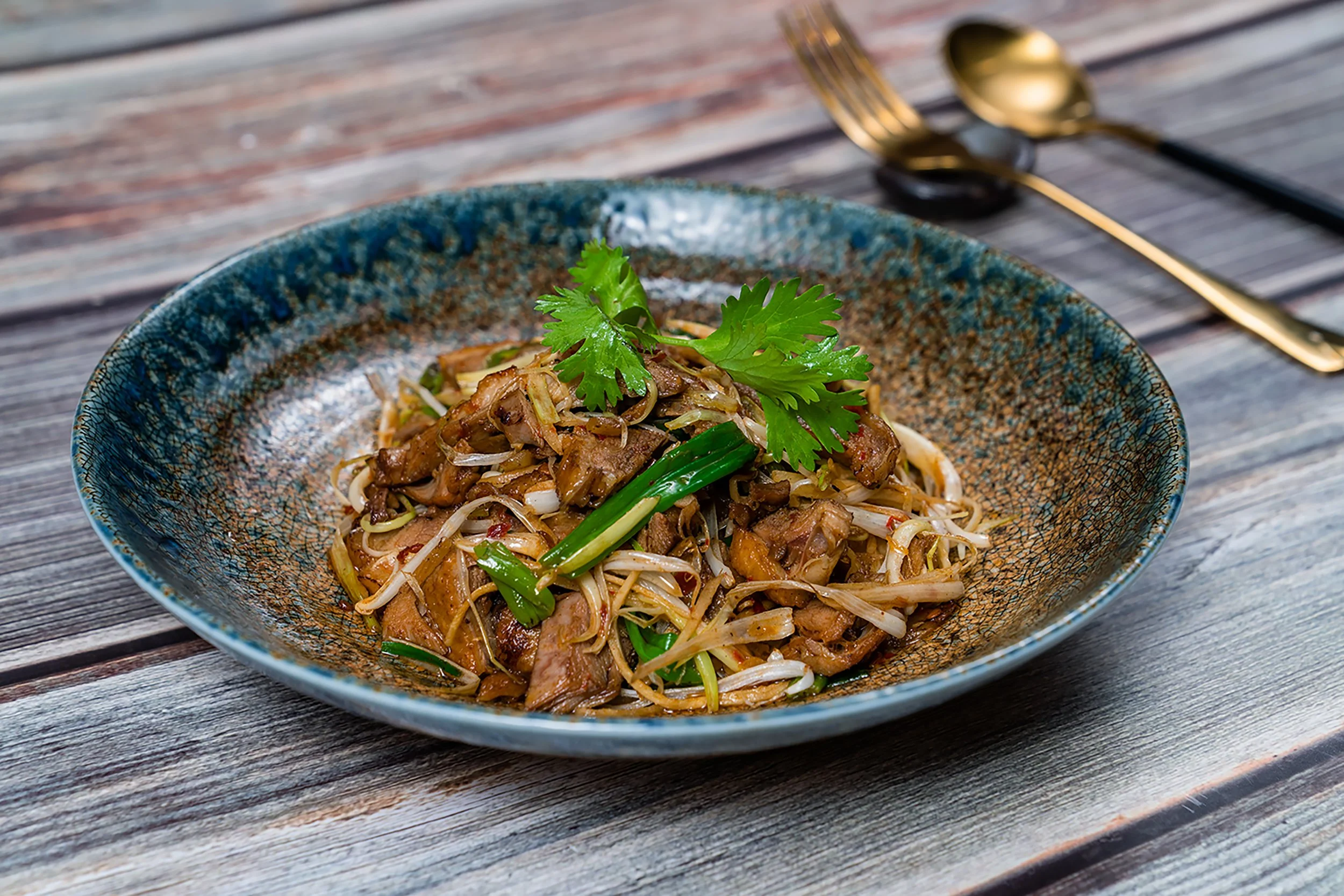 A bowl of Asian-style noodle stir-fry garnished with cilantro on a wooden table with a gold fork and spoon in the background