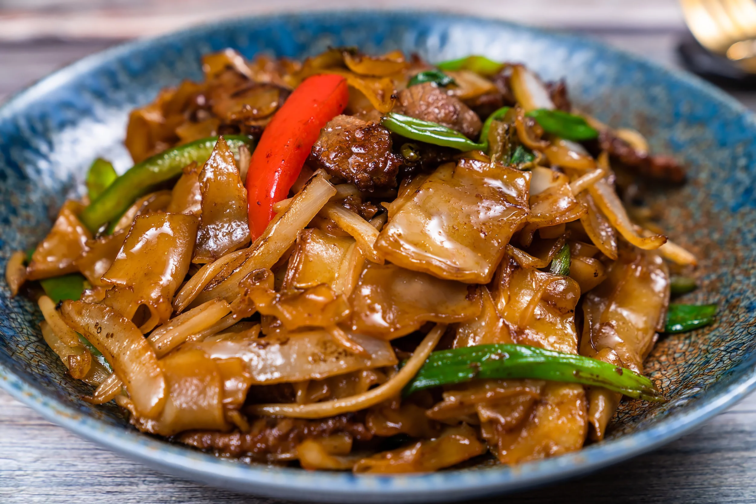 Close-up of a bowl filled with beef stir-fry, including vegetables such as red and green bell peppers, onions, and noodles in a rich brown sauce.