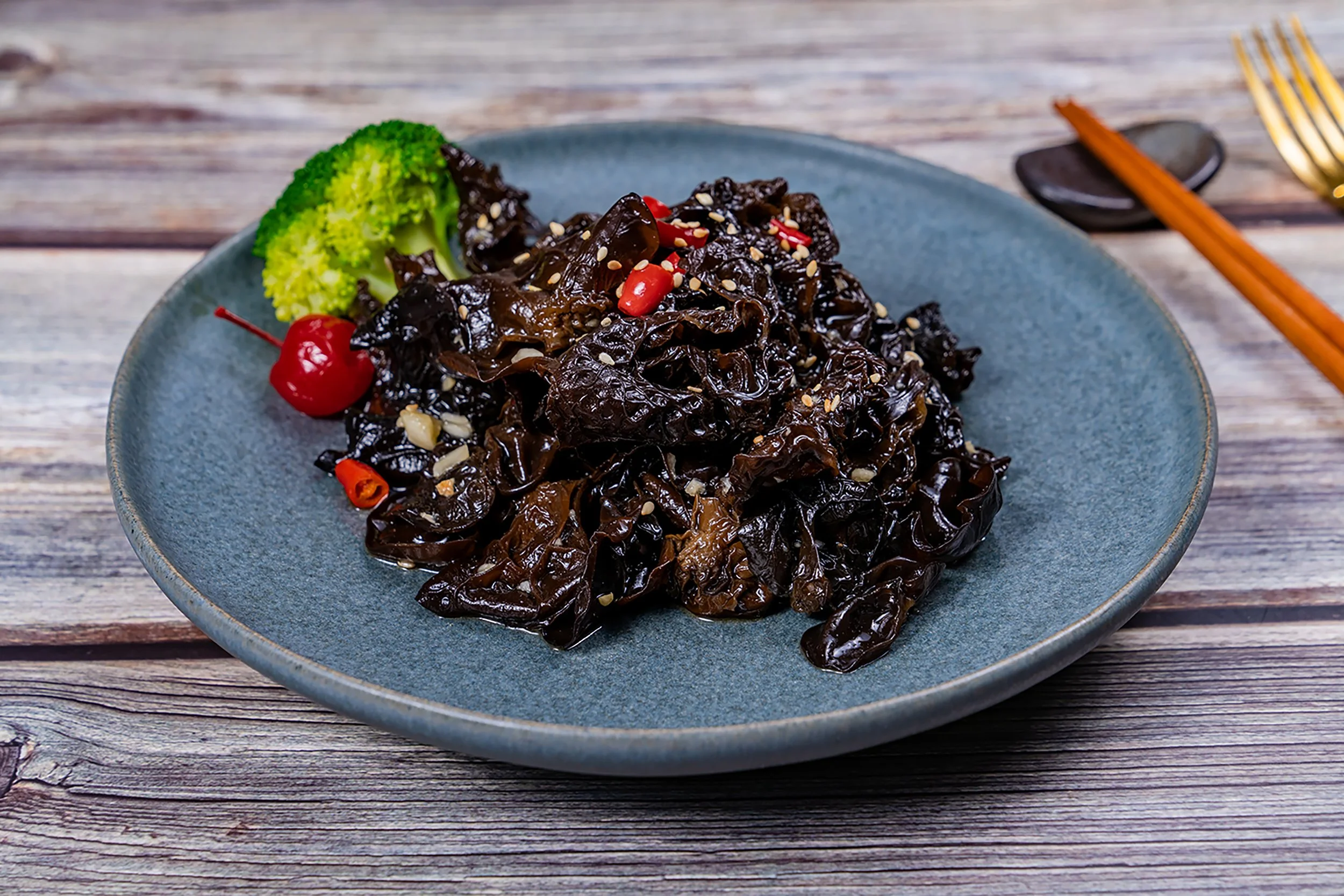 A blue plate with cooked wood ear mushrooms garnished with sesame seeds, sliced red chili peppers, a broccoli floret, and a cherry, placed on a wooden surface.