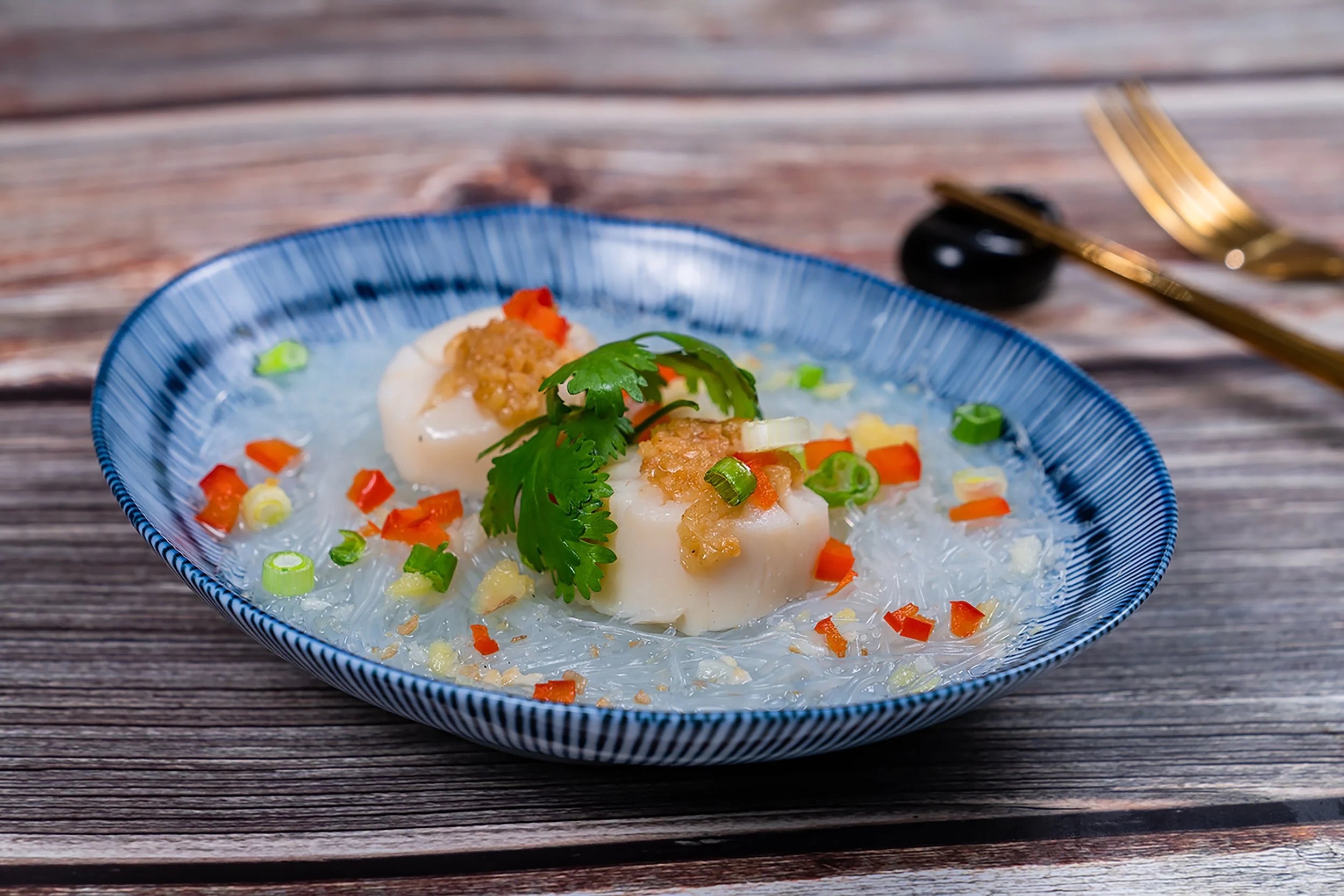 A blue bowl containing a dish with scallops, garnished with cilantro, chopped green onions, diced carrots, and a light broth, placed on a wooden table.