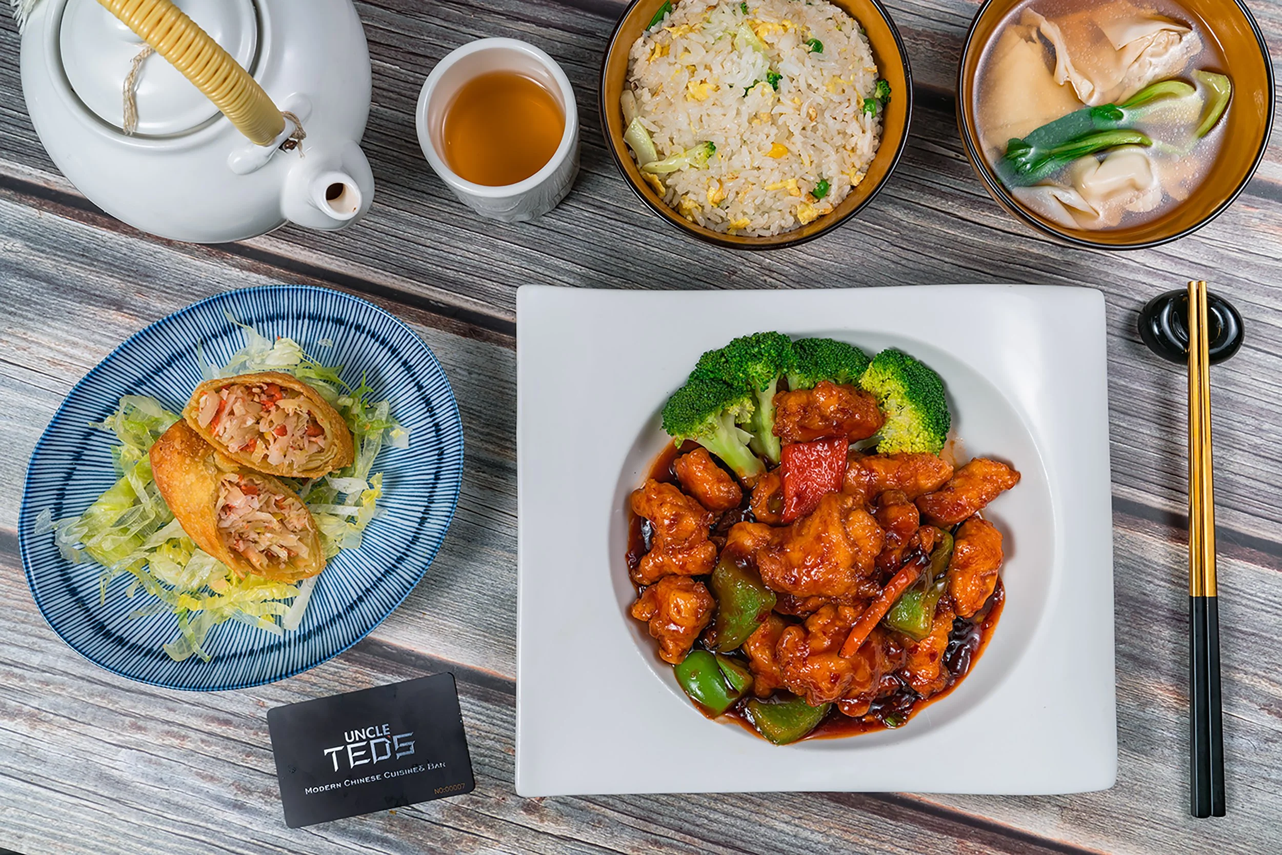 A variety of Chinese dishes on a rustic wooden table, including fried rice with peas and eggs, a bowl of wonton soup with green onions, a spring roll with shredded chicken and vegetables on a bed of lettuce, and a plate of sweet and sour chicken with broccoli and red bell peppers. Chopsticks rest on a black holder, and a tea kettle with a cup of tea is visible in the background.