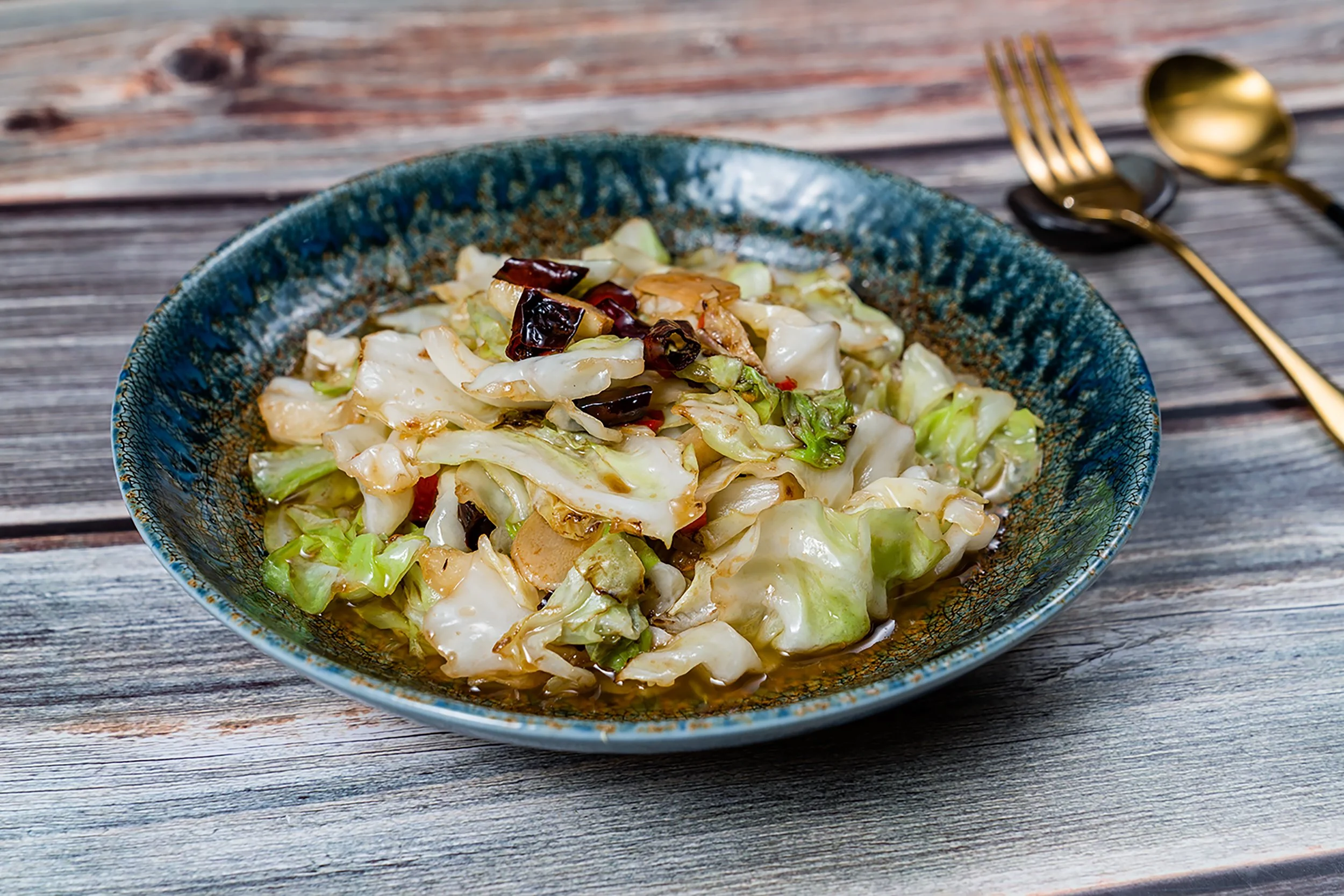 A bowl of stir-fried vegetables with cabbage and dried chili peppers on a wooden table, with a gold spoon and fork in the background.