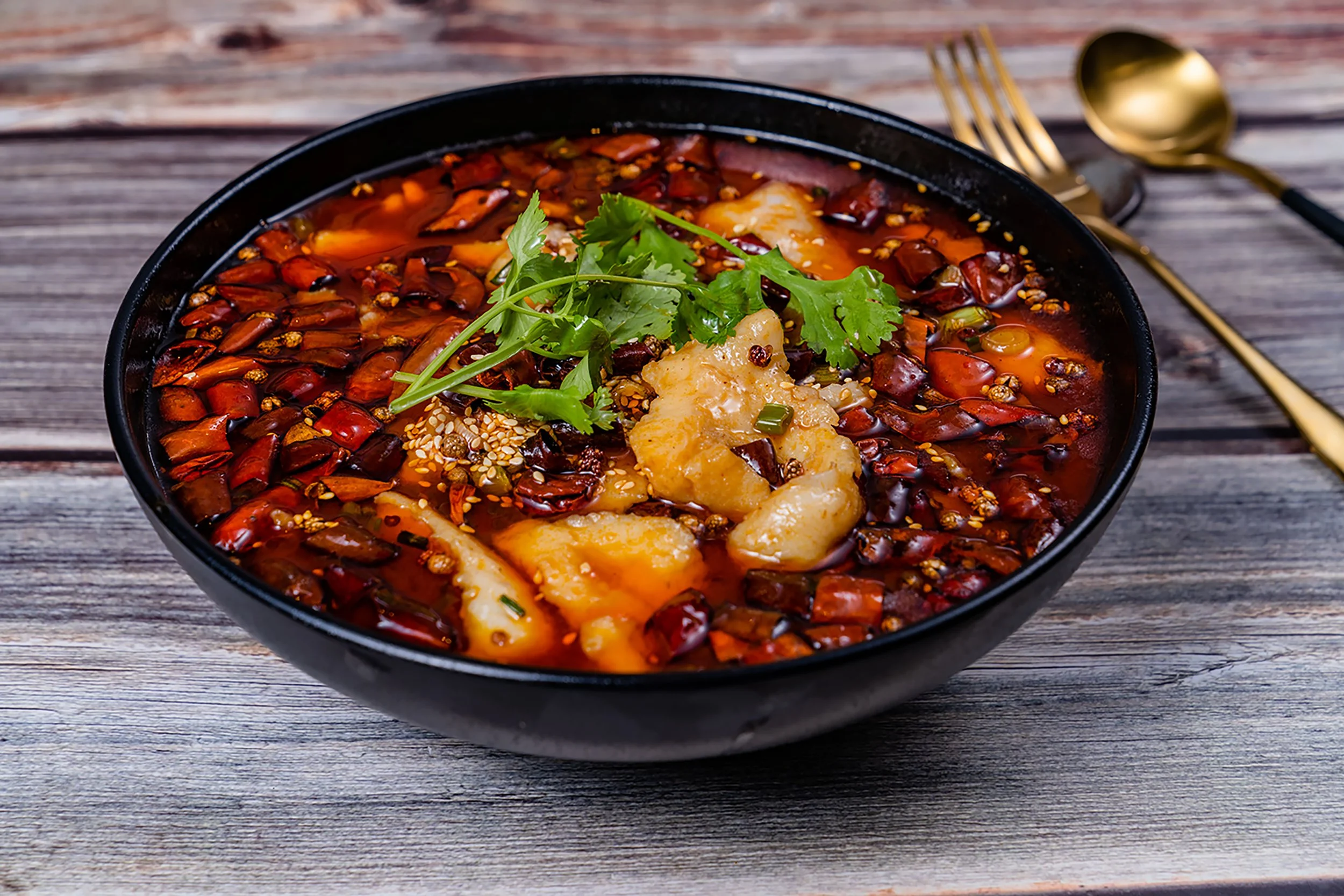 Spicy chili with chunks of potatoes, garnished with cilantro and sesame seeds in a black bowl on a rustic wooden table with gold-colored utensils.