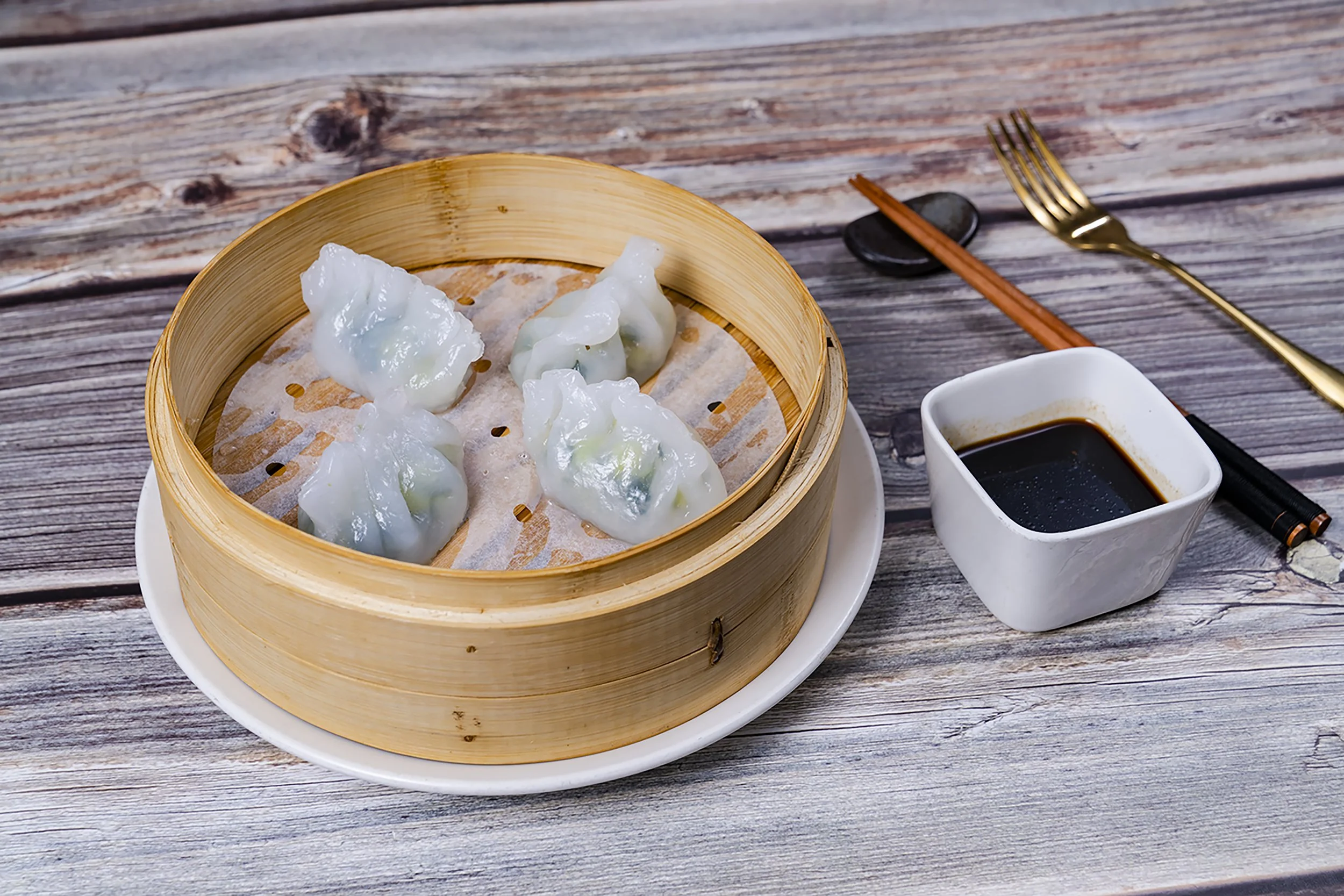 Steamed dumplings in a bamboo basket, on a white plate with soy sauce, chopsticks, a fork, and a small bowl on a weathered wooden table.