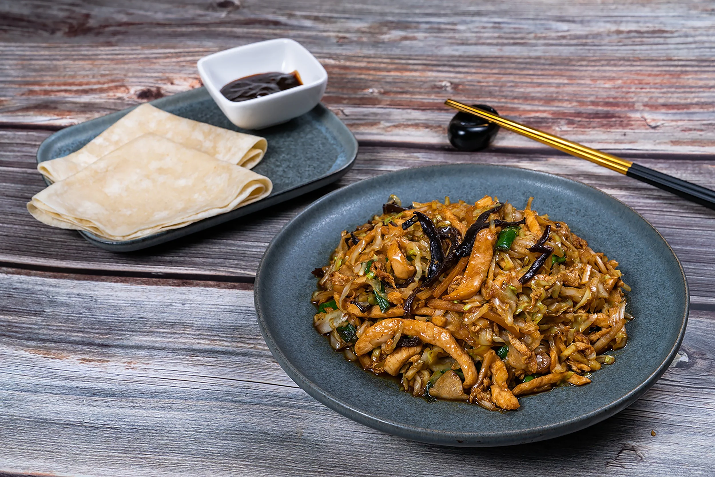 A bowl of Asian stir-fried noodles with vegetables and chicken on a blue plate, set on a rustic wooden table. Next to it, a small dish of soy sauce, a folded napkin, and chopsticks resting on a chopstick holder.