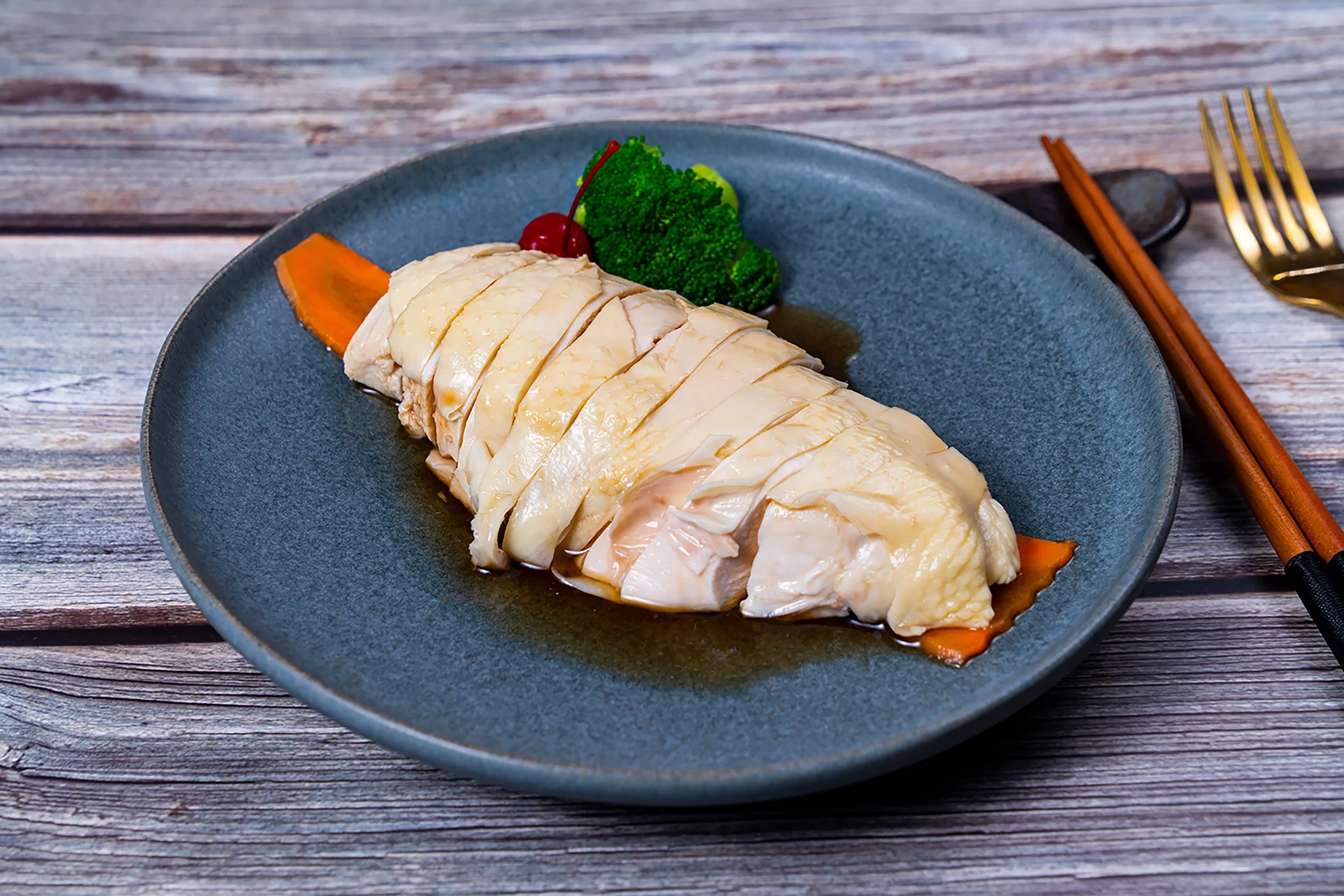 Sliced cooked chicken breast on a blue plate with broccoli, a cherry, and a carrot slice, on a wooden table with chopsticks and a fork.