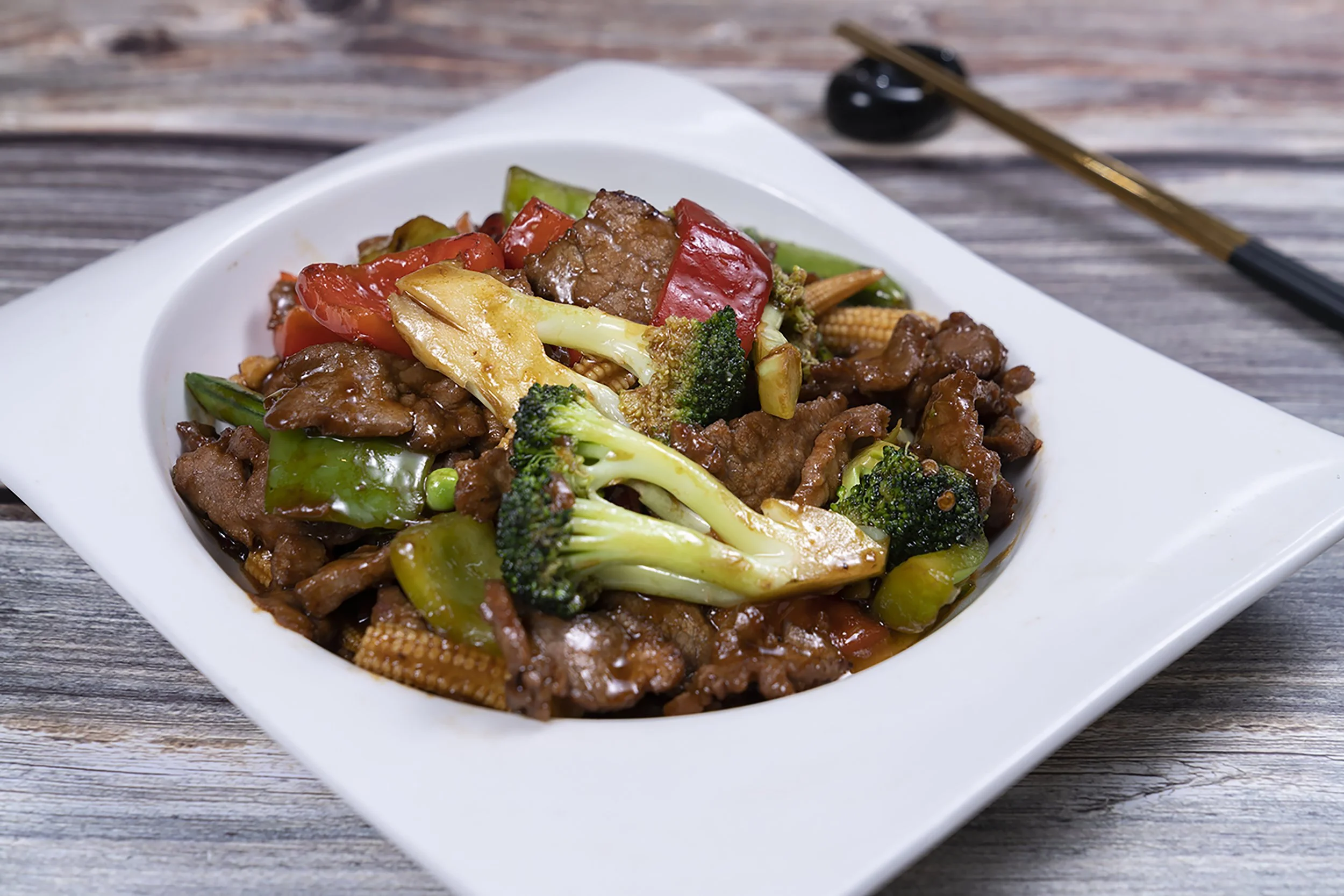 A white rectangular dish contains stir-fried beef and vegetables, including broccoli, red and green bell peppers, baby corn, and carrots, served on a rustic wooden table.