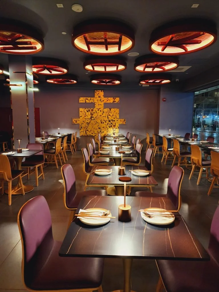 Elegant restaurant interior with dark tables, purple chairs, and a decorative gold wall art piece illuminated by modern lamps with circular and star-like ceiling fixtures.