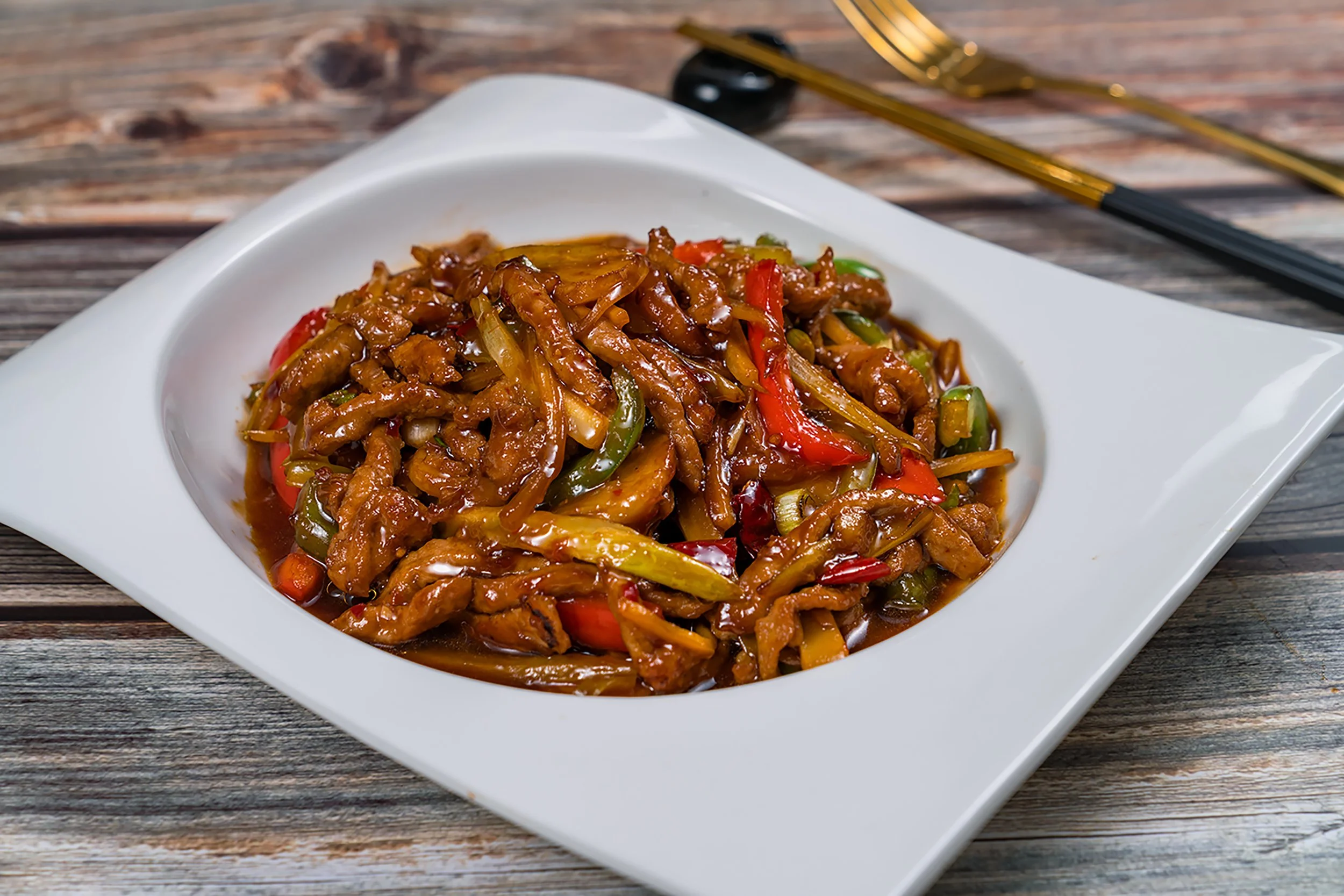 A plate of stir-fried chicken with vegetables in sauce on a white dish, placed on a rustic wooden table with chopsticks in the background.
