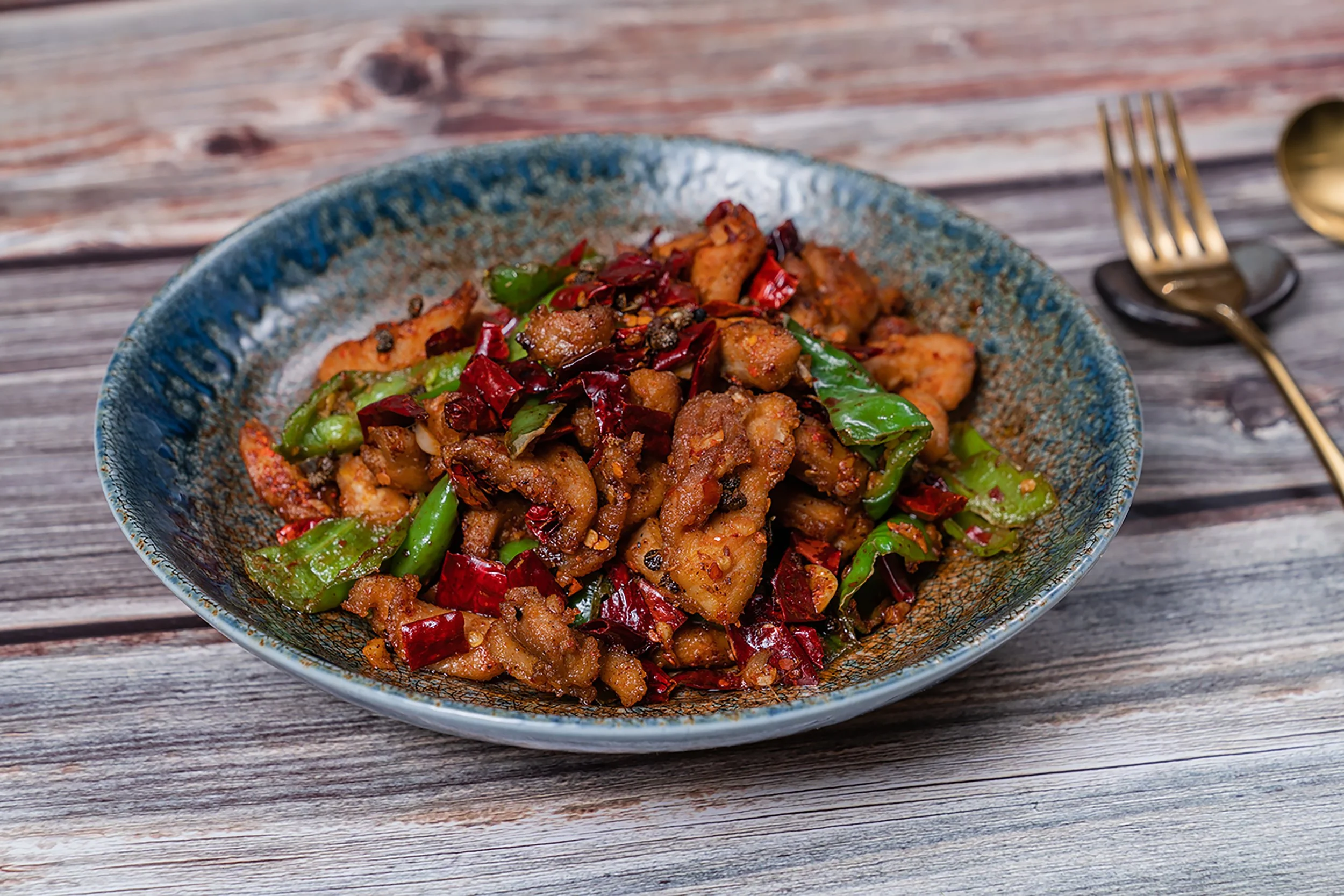A blue ceramic bowl filled with stir-fried chicken, red and green chili peppers, and sliced vegetables on a wooden table with a gold-handled fork and spoon.